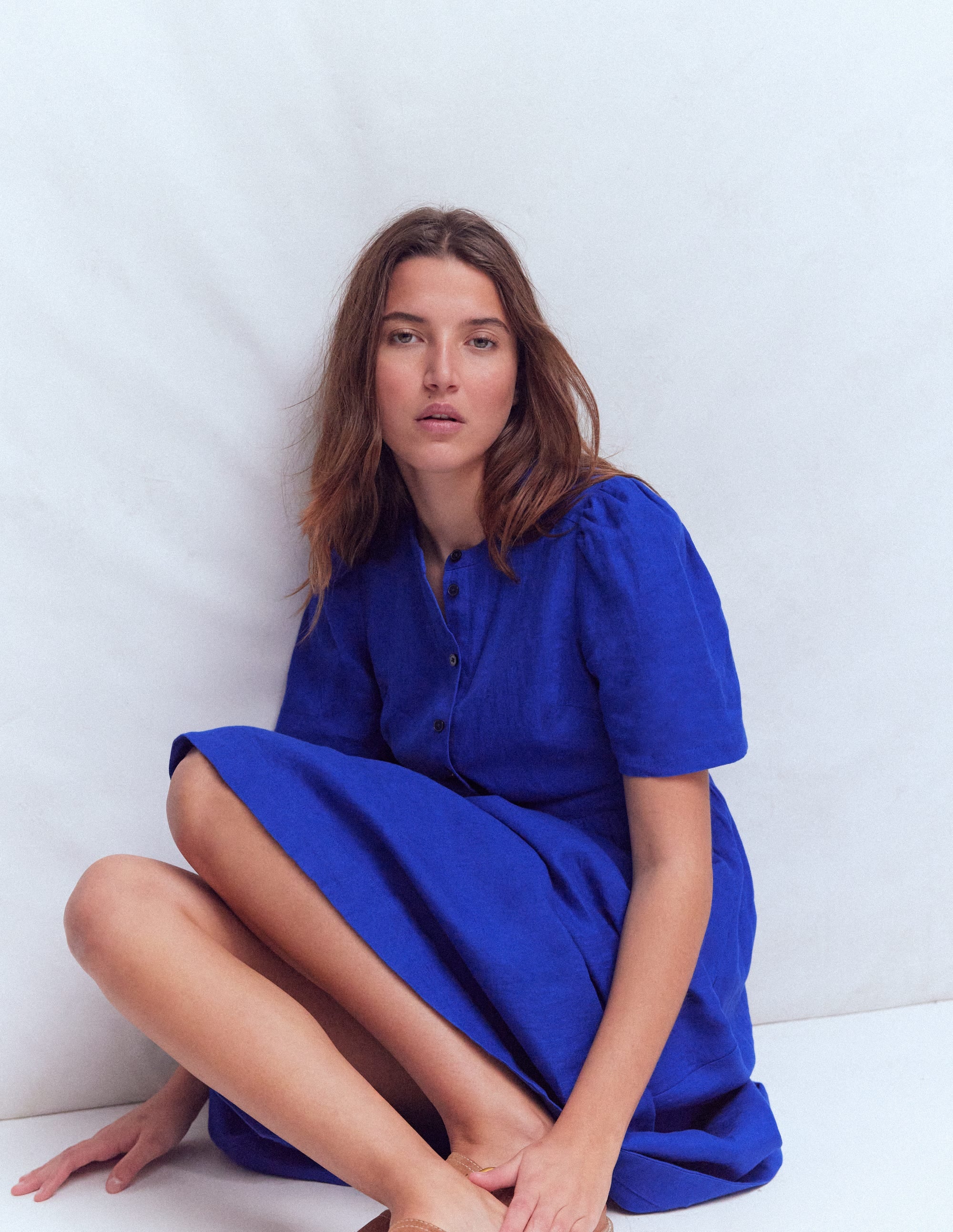 A woman wearing a bright blue linen midi dress with a wide waistband and an open button placket at the neck, sitting against a white background.