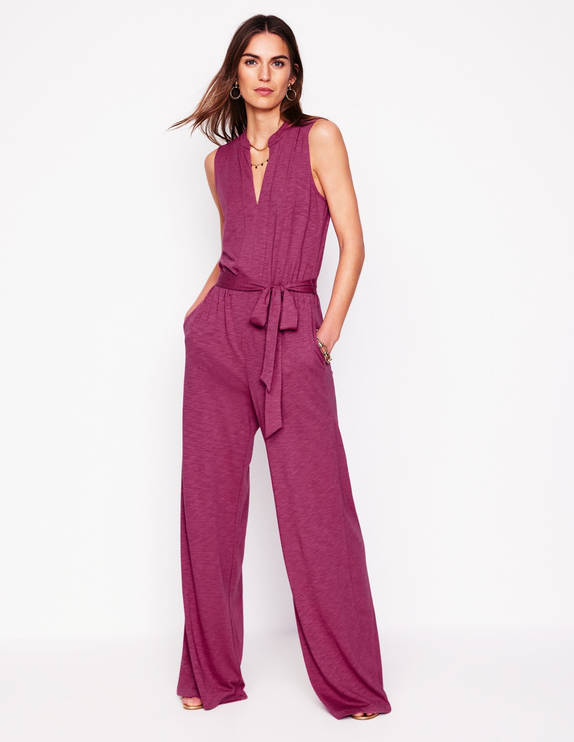 A woman wearing a plum purple jumpsuit with a notch neckline, wide legs, and a self-tie belt, standing against a white background.