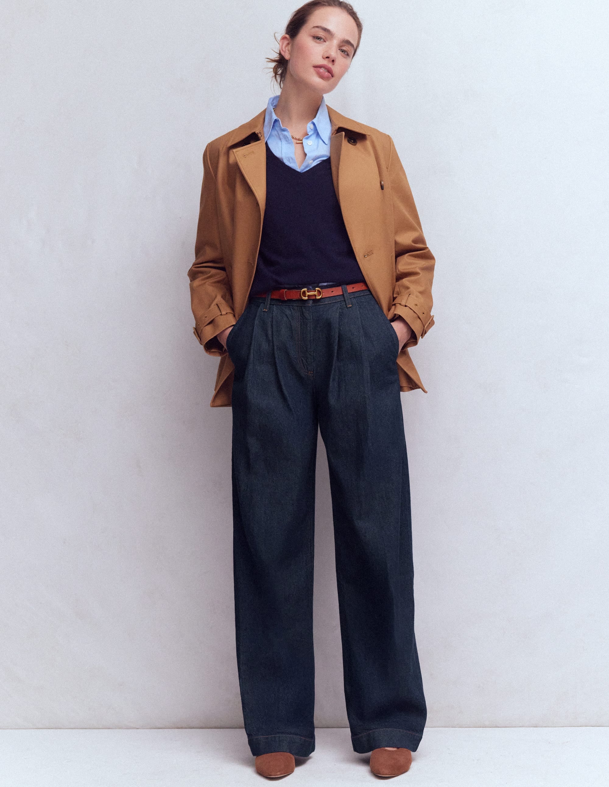 A woman wearing indigo rinse pleated wide-leg jeans with a high waist, paired with a blue shirt, navy sweater, and tan belt.