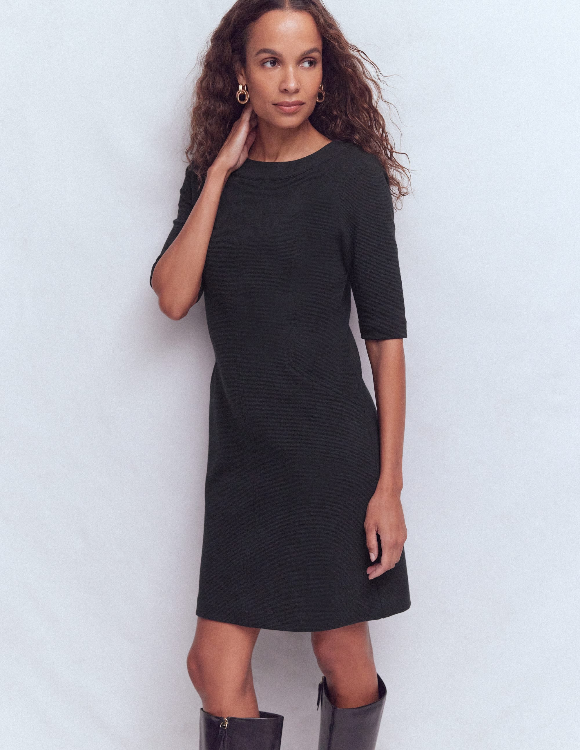 A woman wearing a black shift dress with a round neckline, three-quarter sleeves, and side seam pockets, standing against a plain background.