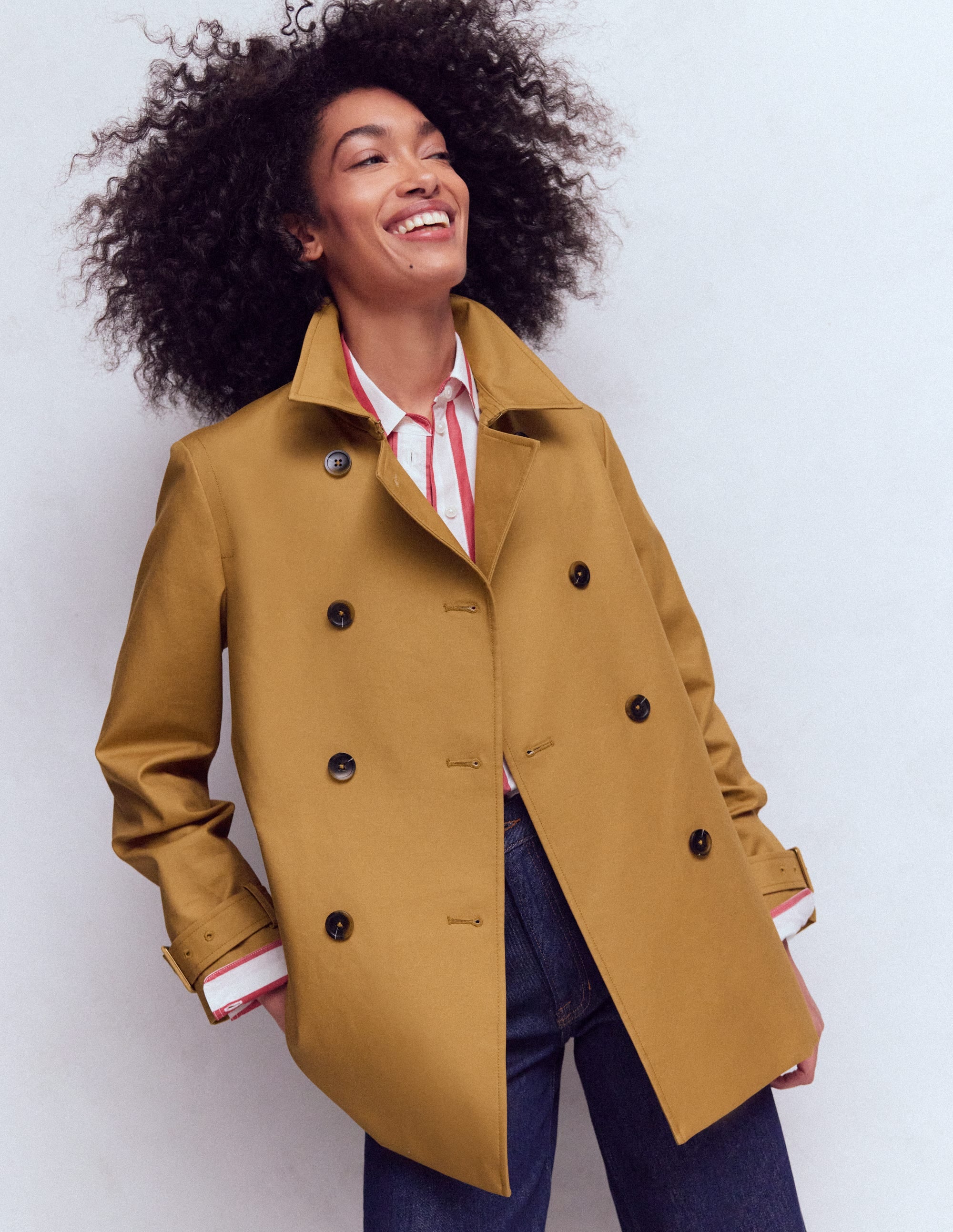 A woman wearing a mustard yellow swing trench coat with a wide collar, black buttons, and functional pockets, standing against a plain background.