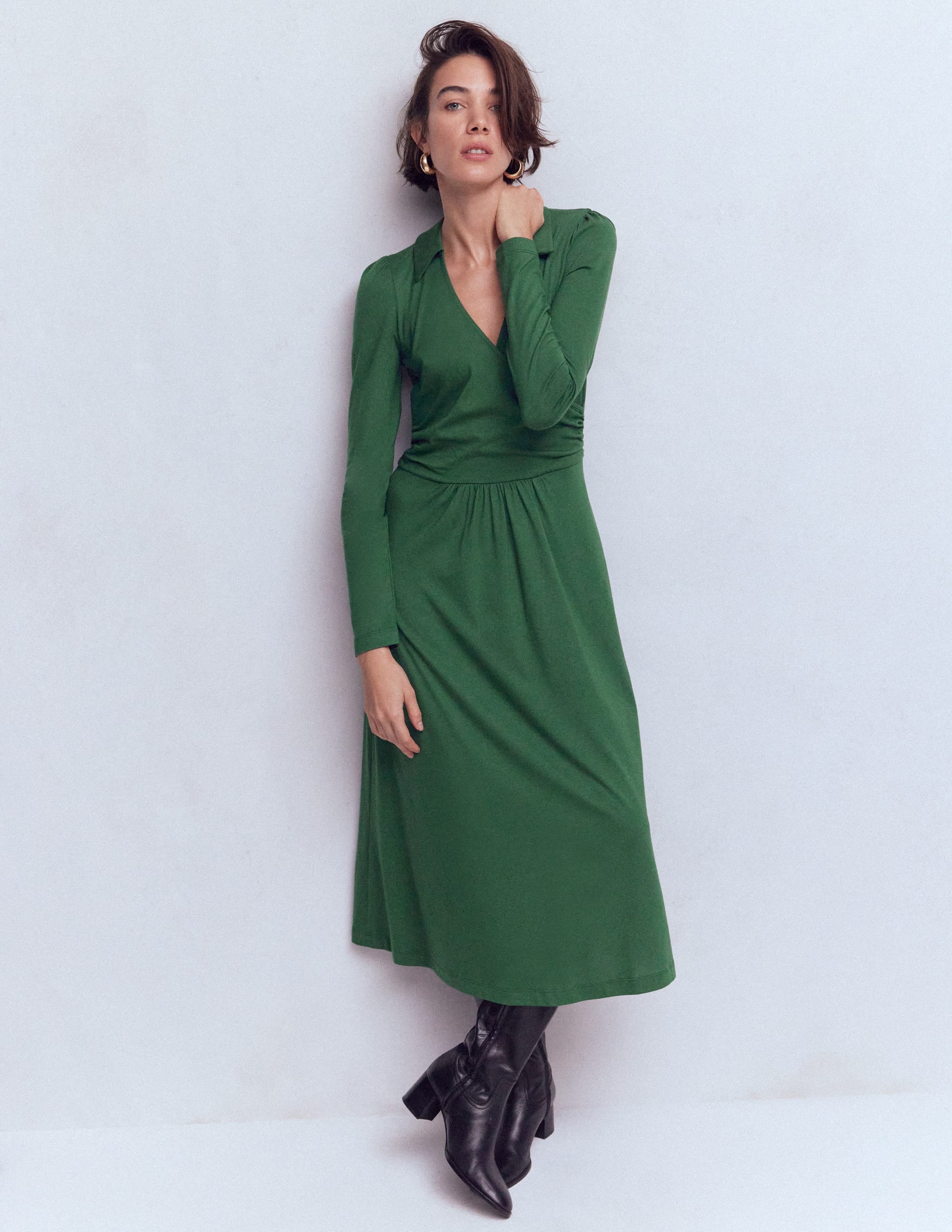 A woman wearing a green wrap dress with long sleeves, a V-neckline, and a gathered waist, paired with black knee-high boots.