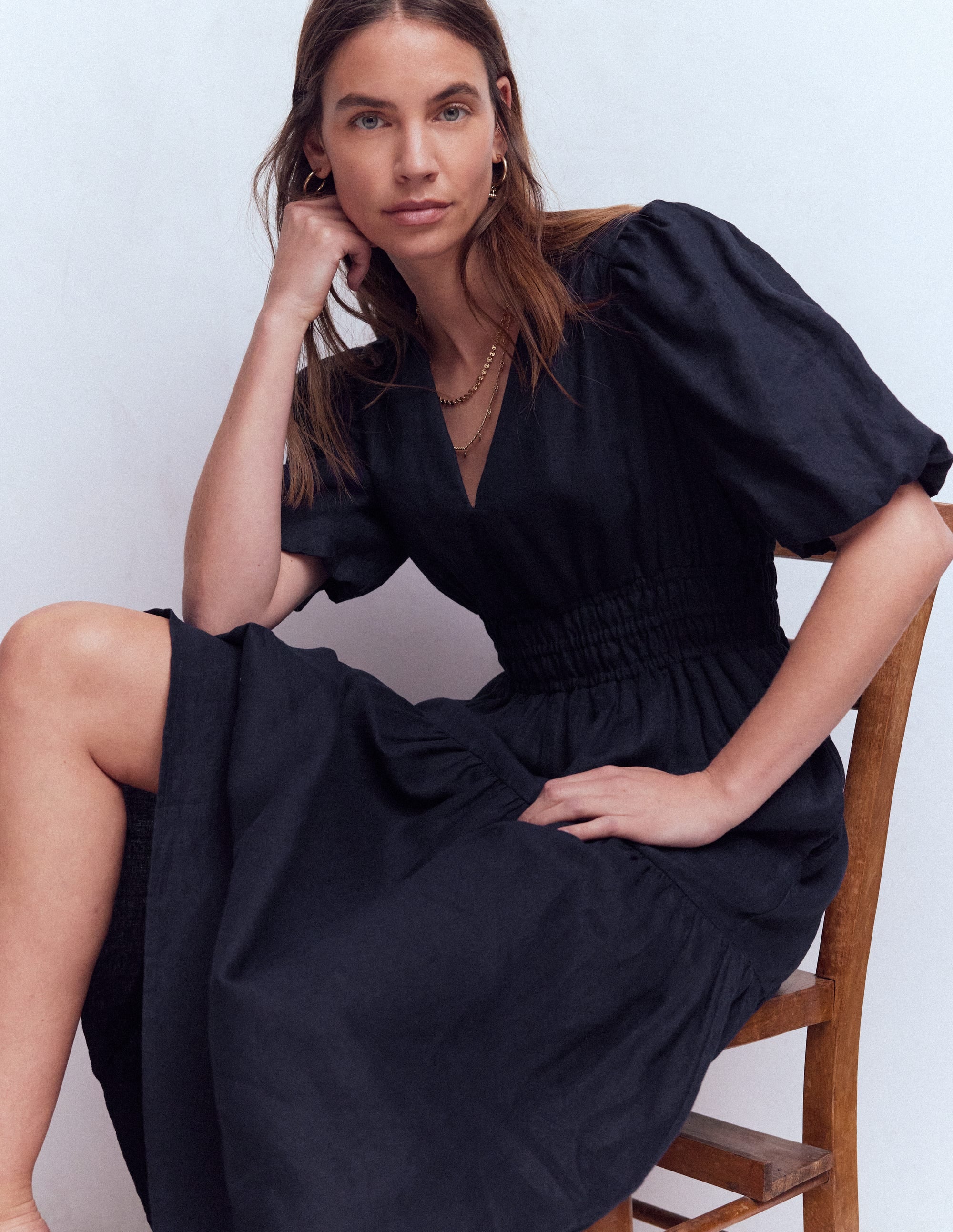 A woman wearing a navy linen dress with puff sleeves, fitted waist, and maxi length, sitting on a wooden chair.