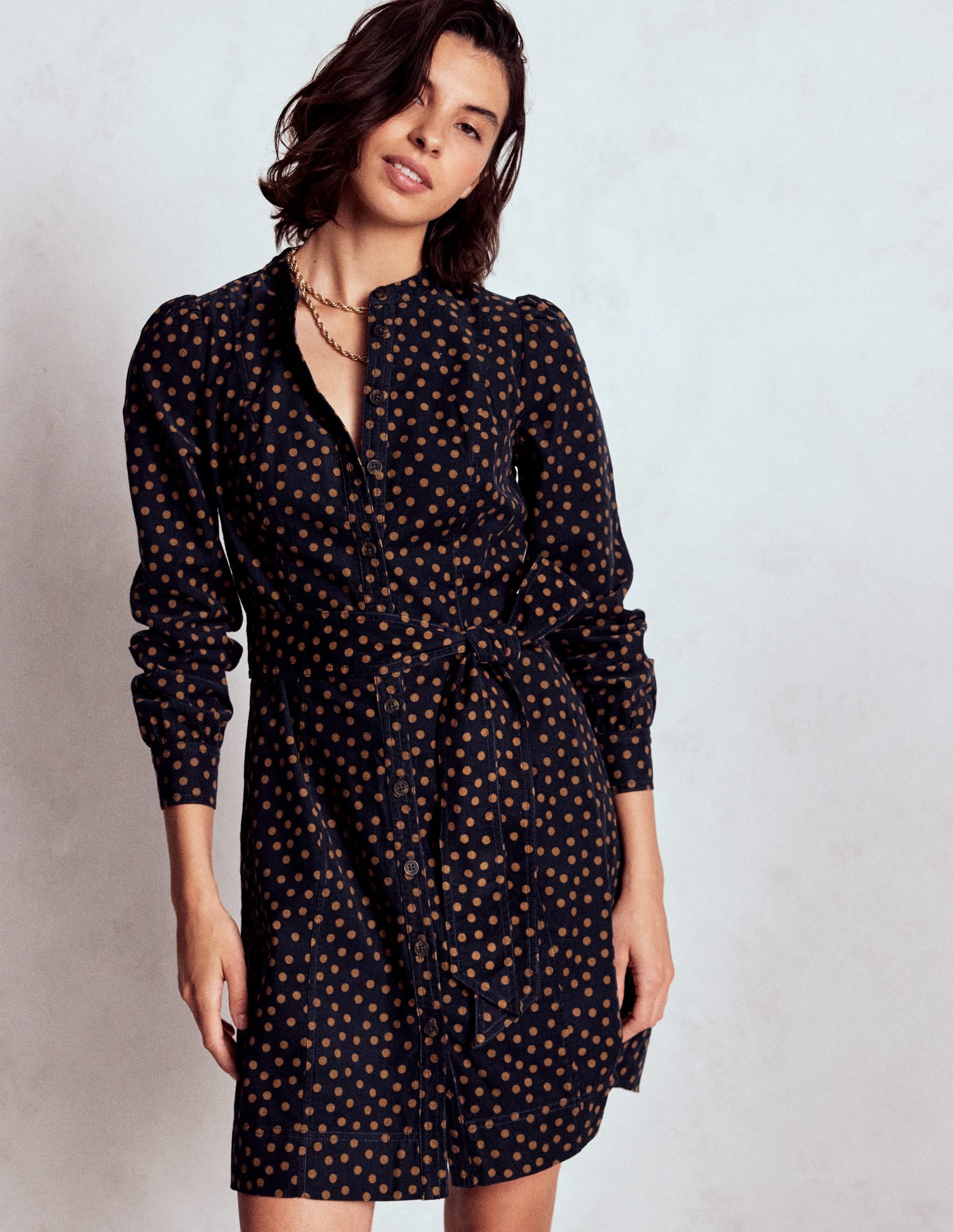 A woman wearing a navy blue short dress with orange painted dots, long sleeves, and a fabric belt, standing against a plain background.