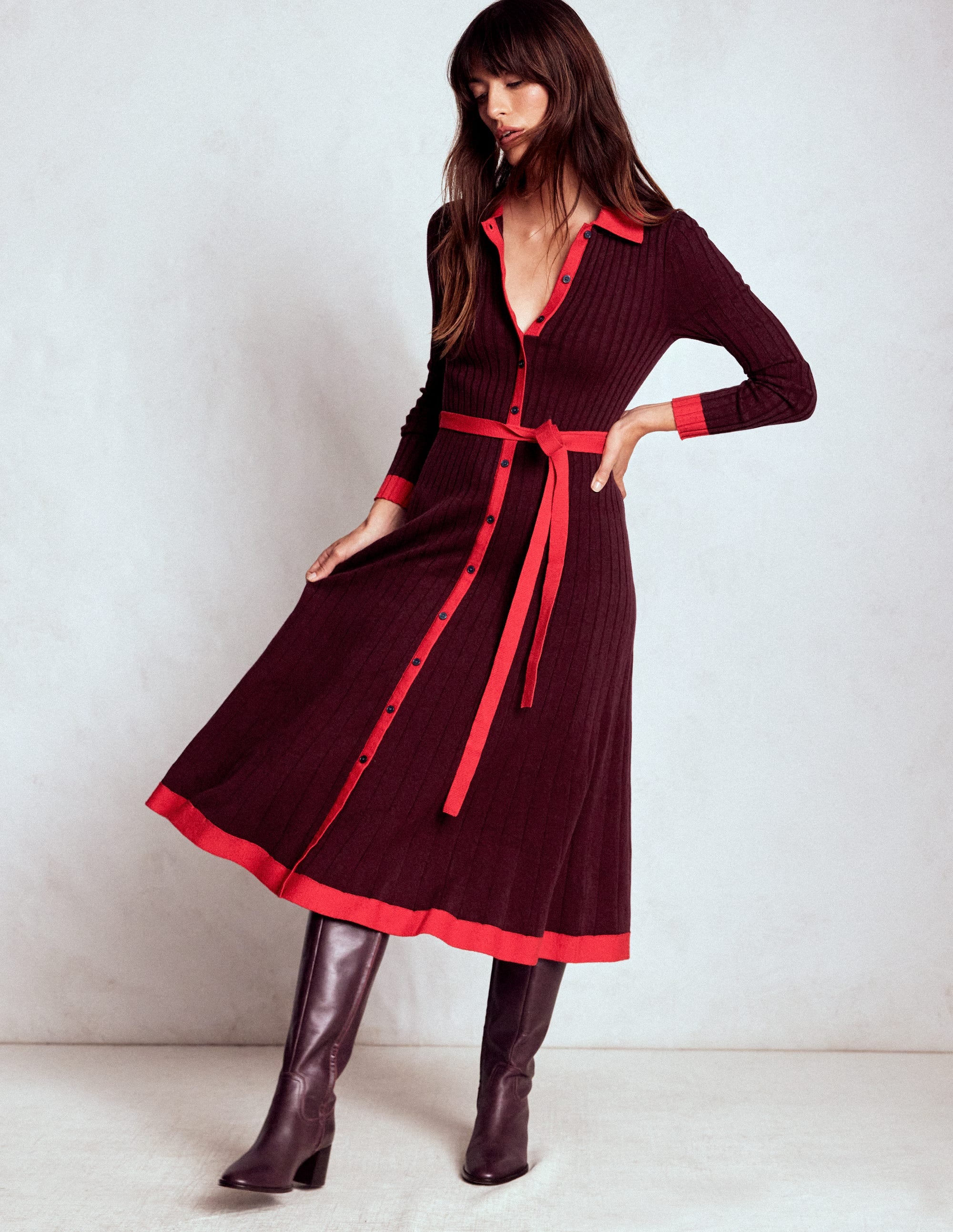 A woman wearing a deep wisteria knitted shirt dress with red accents, a belt, and knee-high boots.