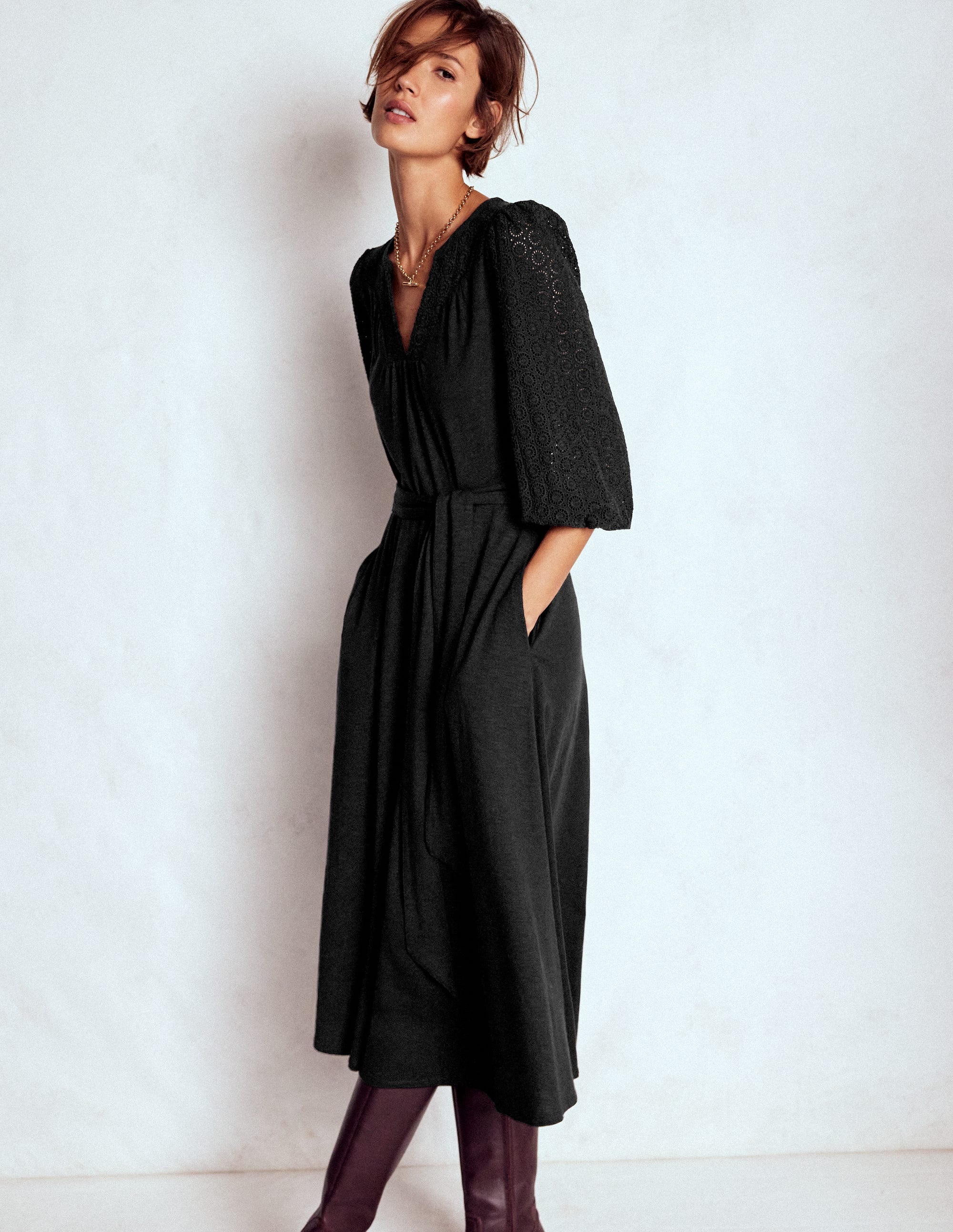 A woman wearing a black Eden Broderie Jersey Dress with embroidered sleeves, V-neckline, and a self-fabric belt, standing against a plain background.