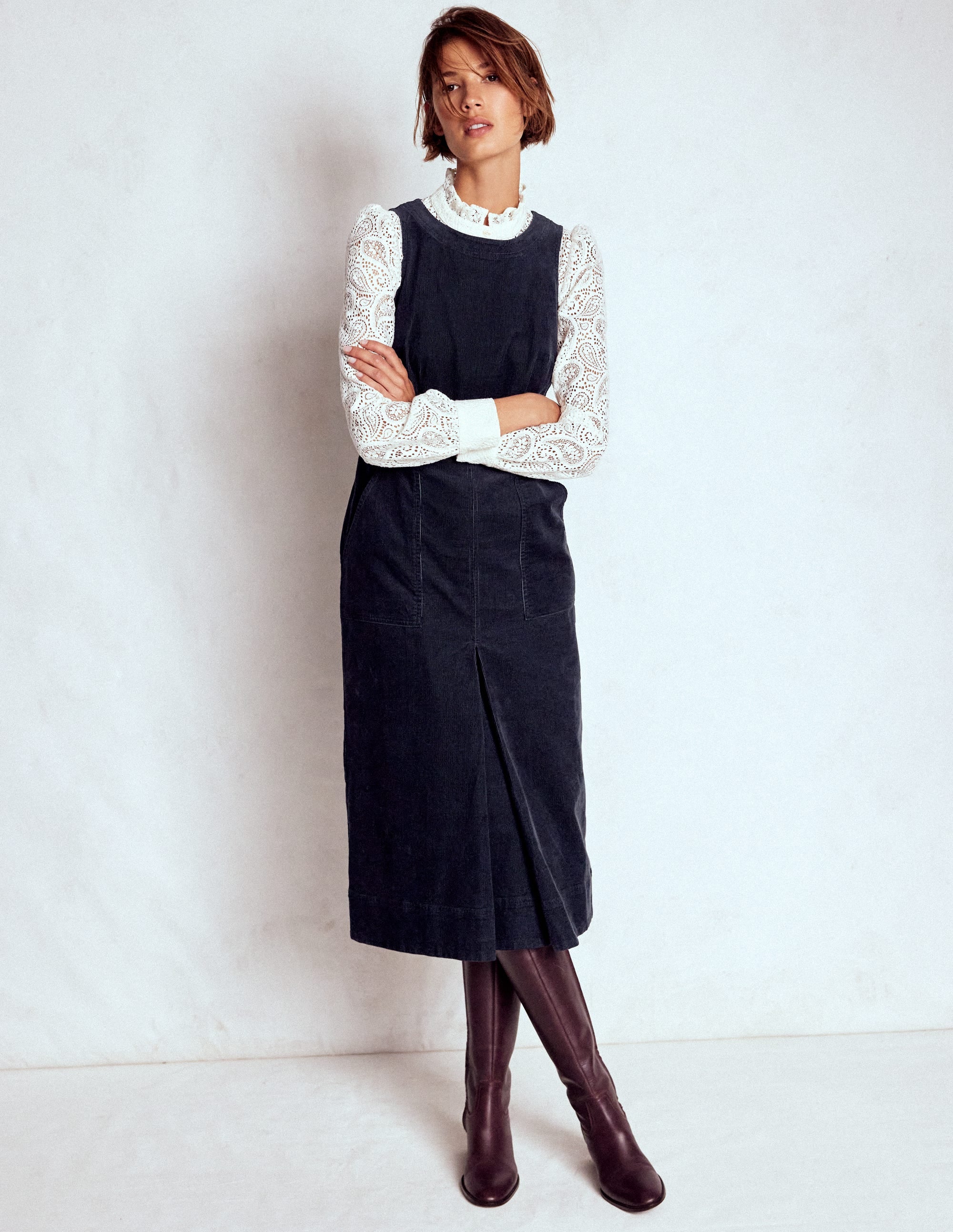 A woman wearing a midnight blue midi dress with a box pleat design, paired with a white lace long-sleeve top and brown knee-high boots.