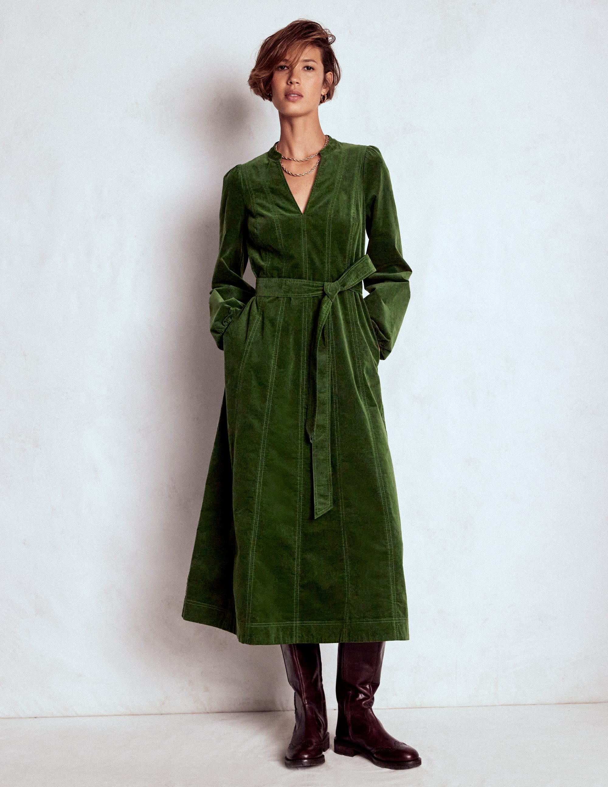 A woman wearing a green midi dress with long sleeves, a self-fabric belt, and pockets, paired with dark boots.