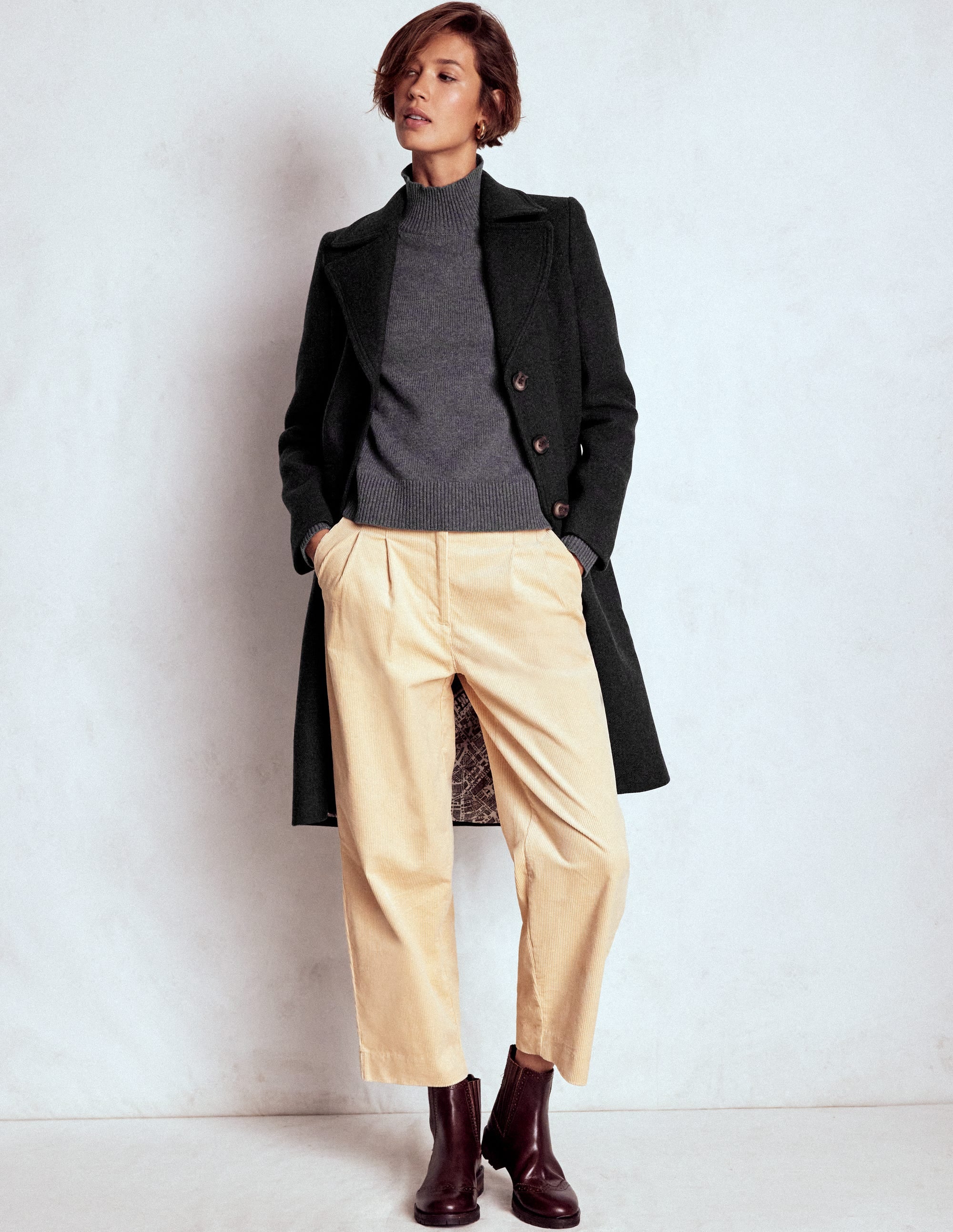 A woman wearing a black wool coat with a button front, over a grey turtleneck sweater, beige trousers, and dark brown boots.