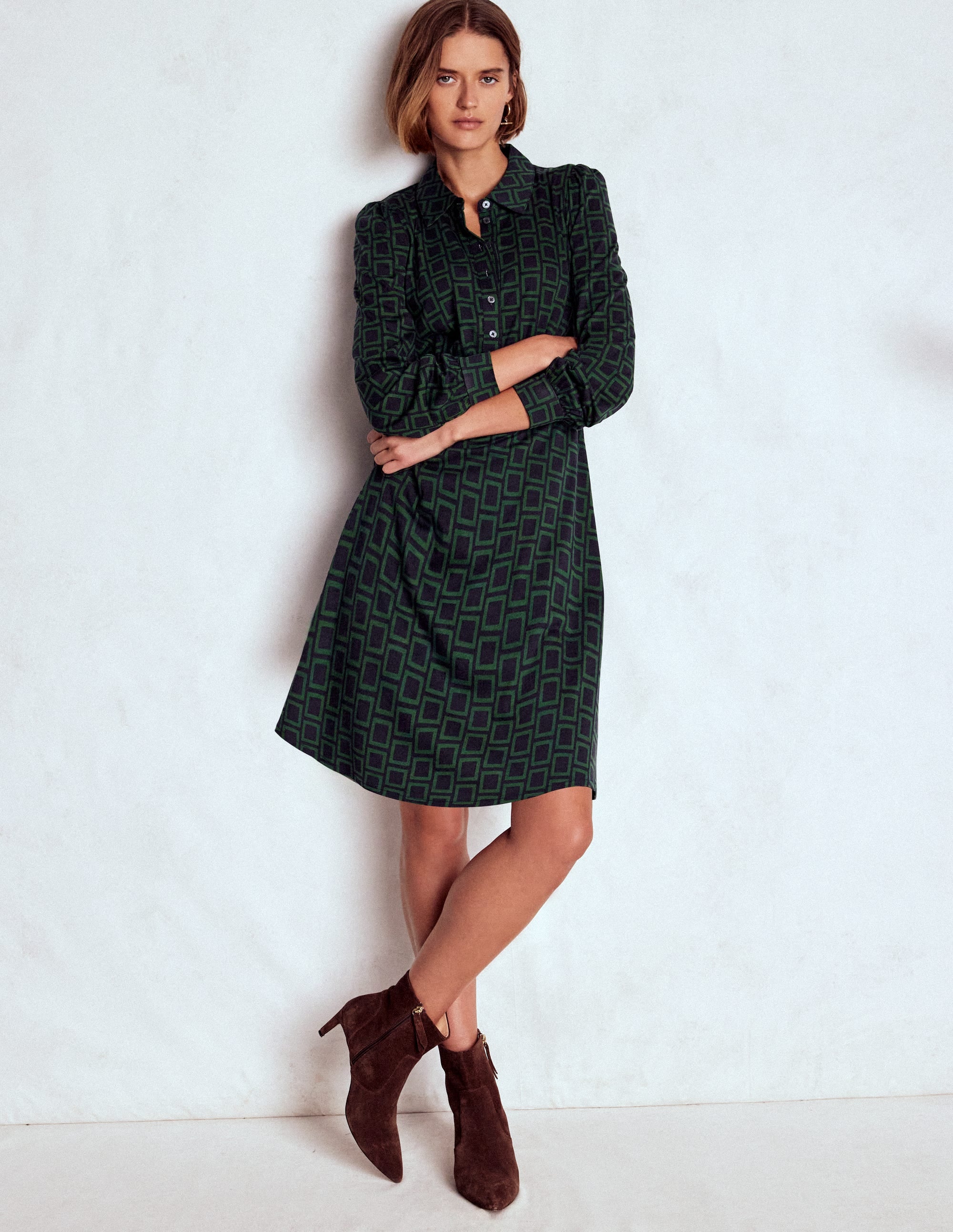 A woman wearing a dark green shirt dress with a square geometric pattern, long sleeves, collar, and button-down front, standing against a plain background.