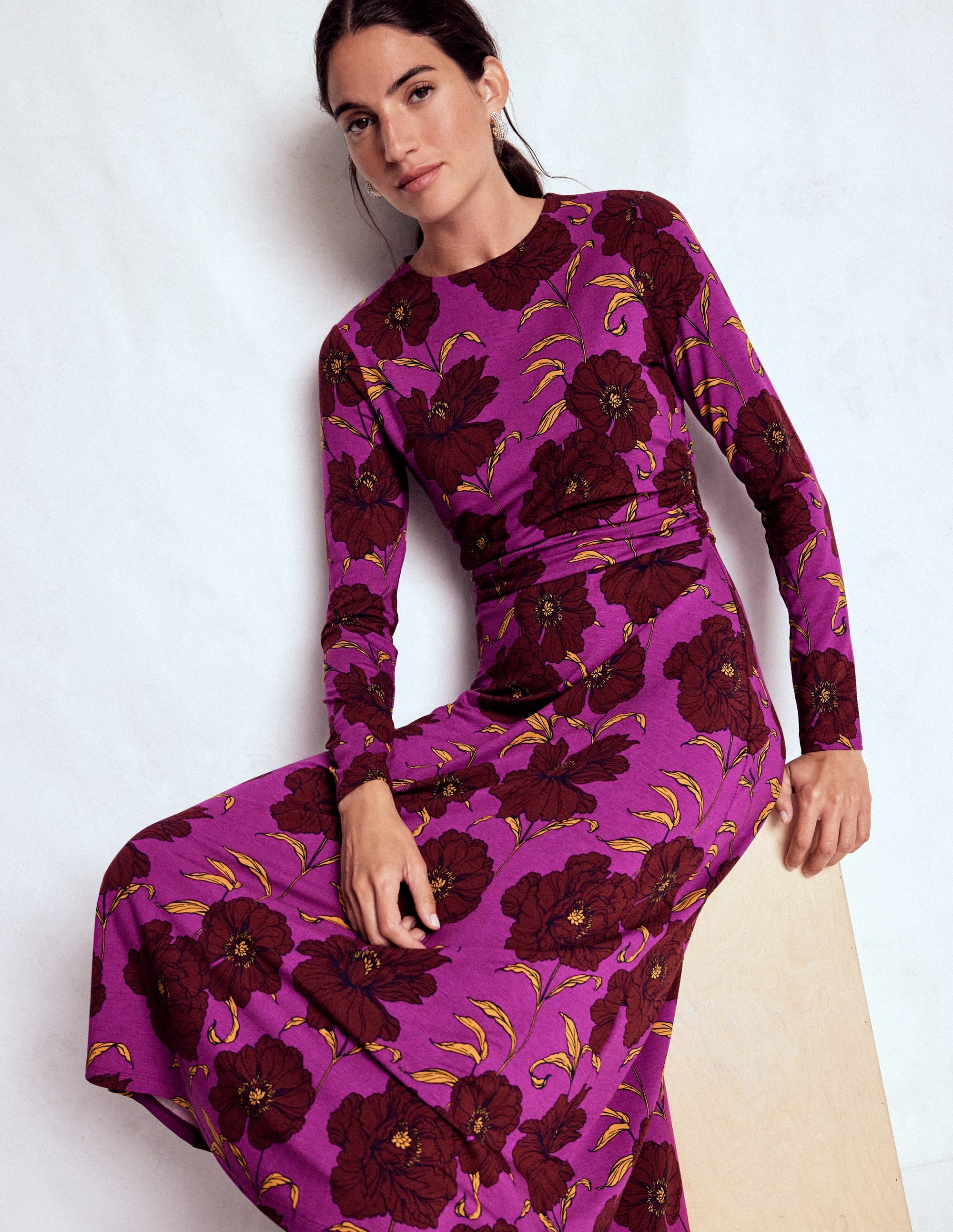A woman wearing a purple floral midi dress with long sleeves and ruched detailing, featuring a deep red and yellow floral pattern.