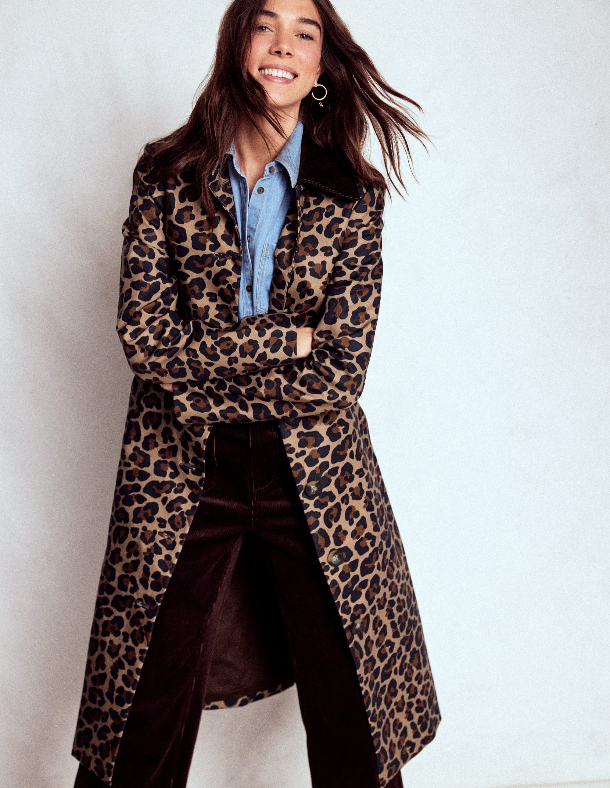 A woman wearing a long leopard print coat with a belt, black collar, and pockets, over a blue shirt, smiling.