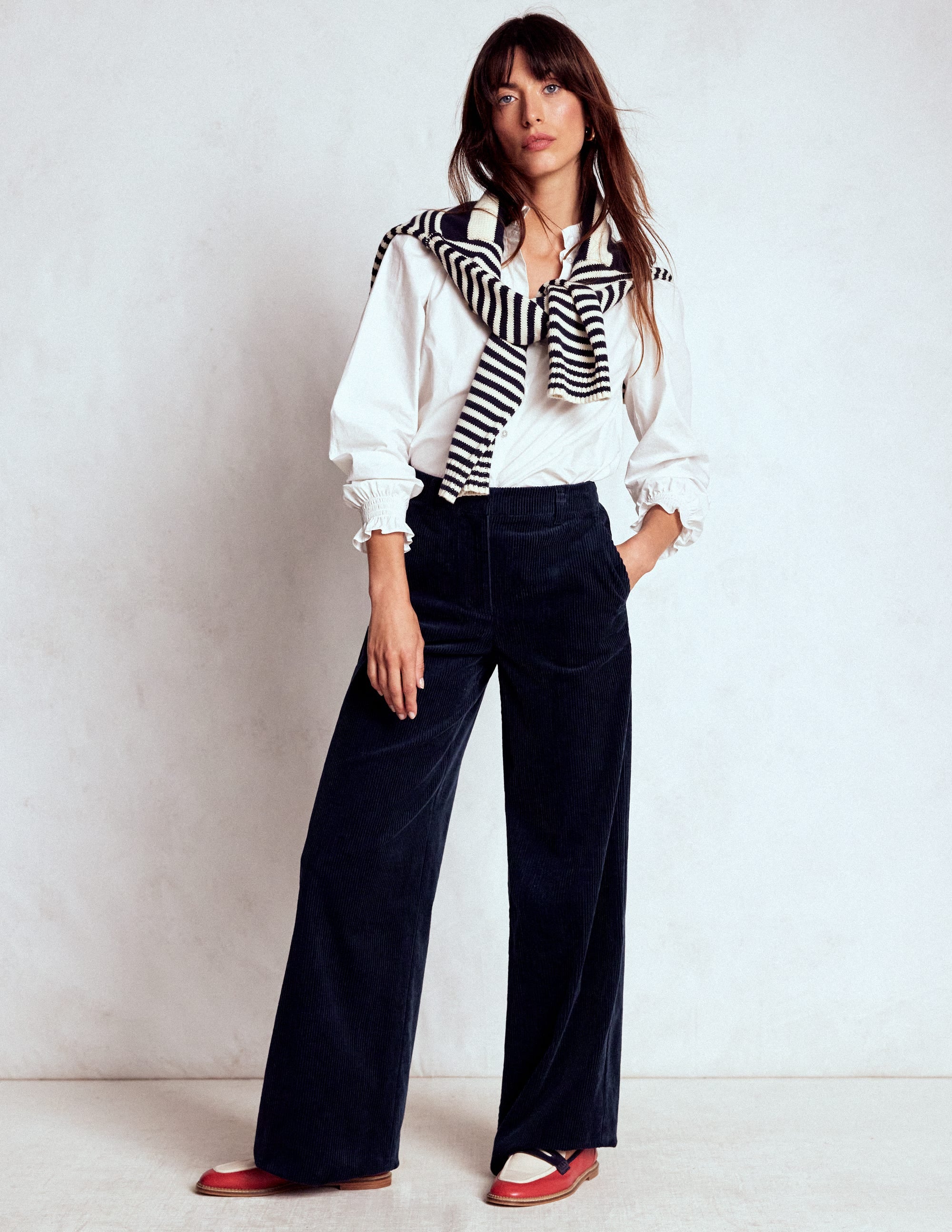 A woman wearing navy wide-leg cord trousers, a white blouse, and a striped sweater draped over her shoulders.