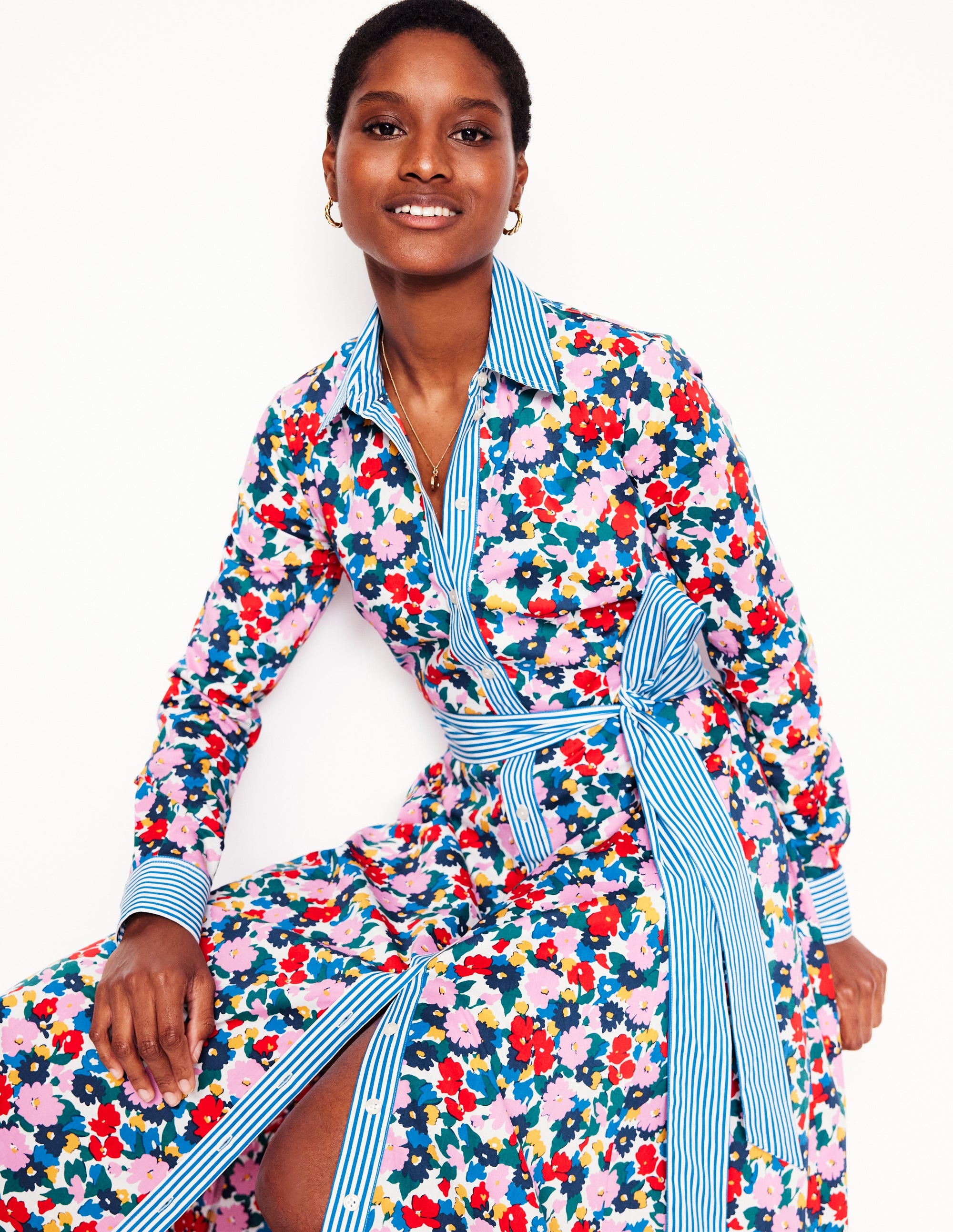 A woman wearing a colorful floral midi shirt dress with a blue and white striped collar and belt, featuring a button-down front, long sleeves with cuffs, and side pockets.