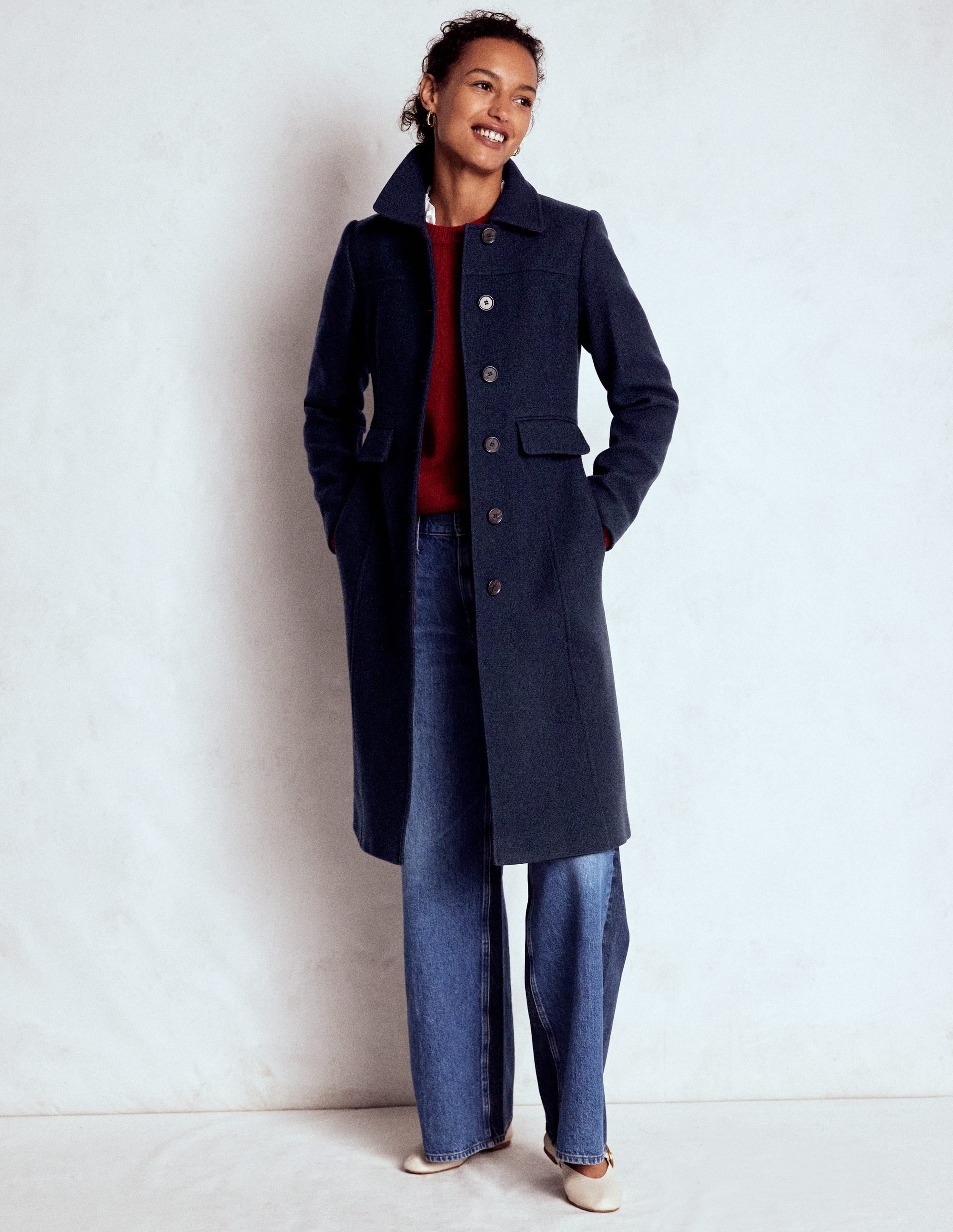 A woman wearing a navy blue fitted coat with a classic collar, button front, and knee-length cut, paired with jeans and white shoes.