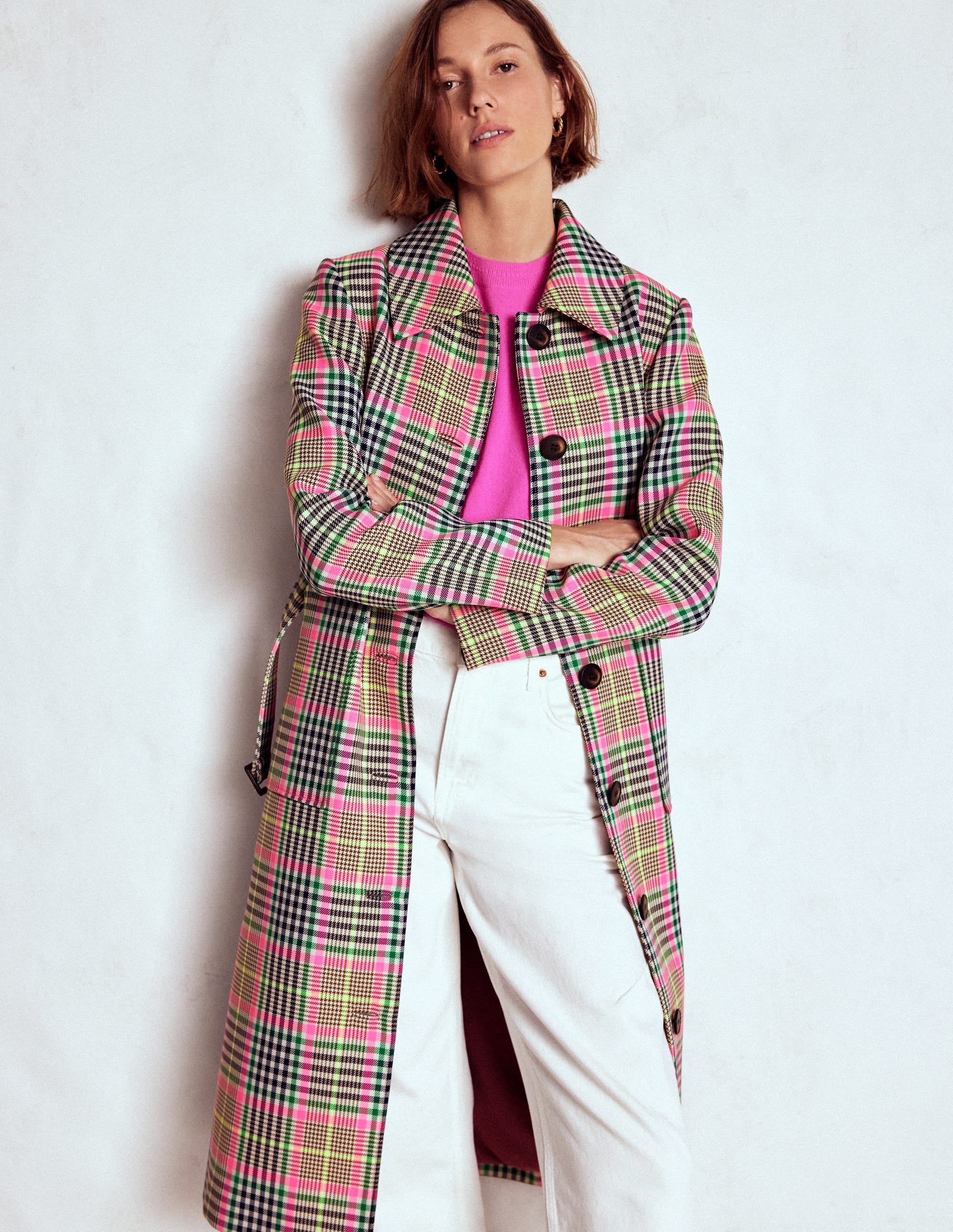 A woman wearing a belted mac coat with a brown and neon pink check pattern, buttoned at the front, with pockets and a detachable waist tie.