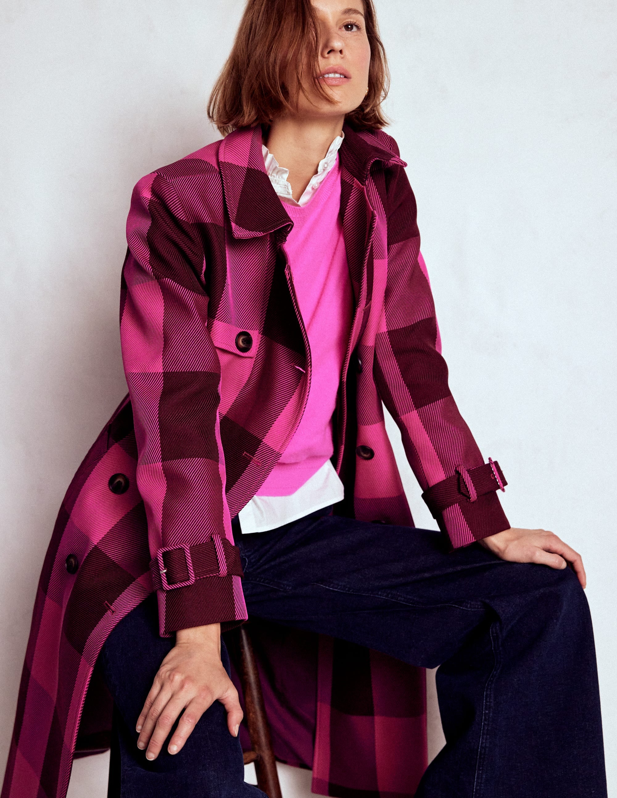 A woman wearing a pink and chestnut gingham patterned trench coat with a double-breasted front, waist belt, and pockets, sitting on a chair.