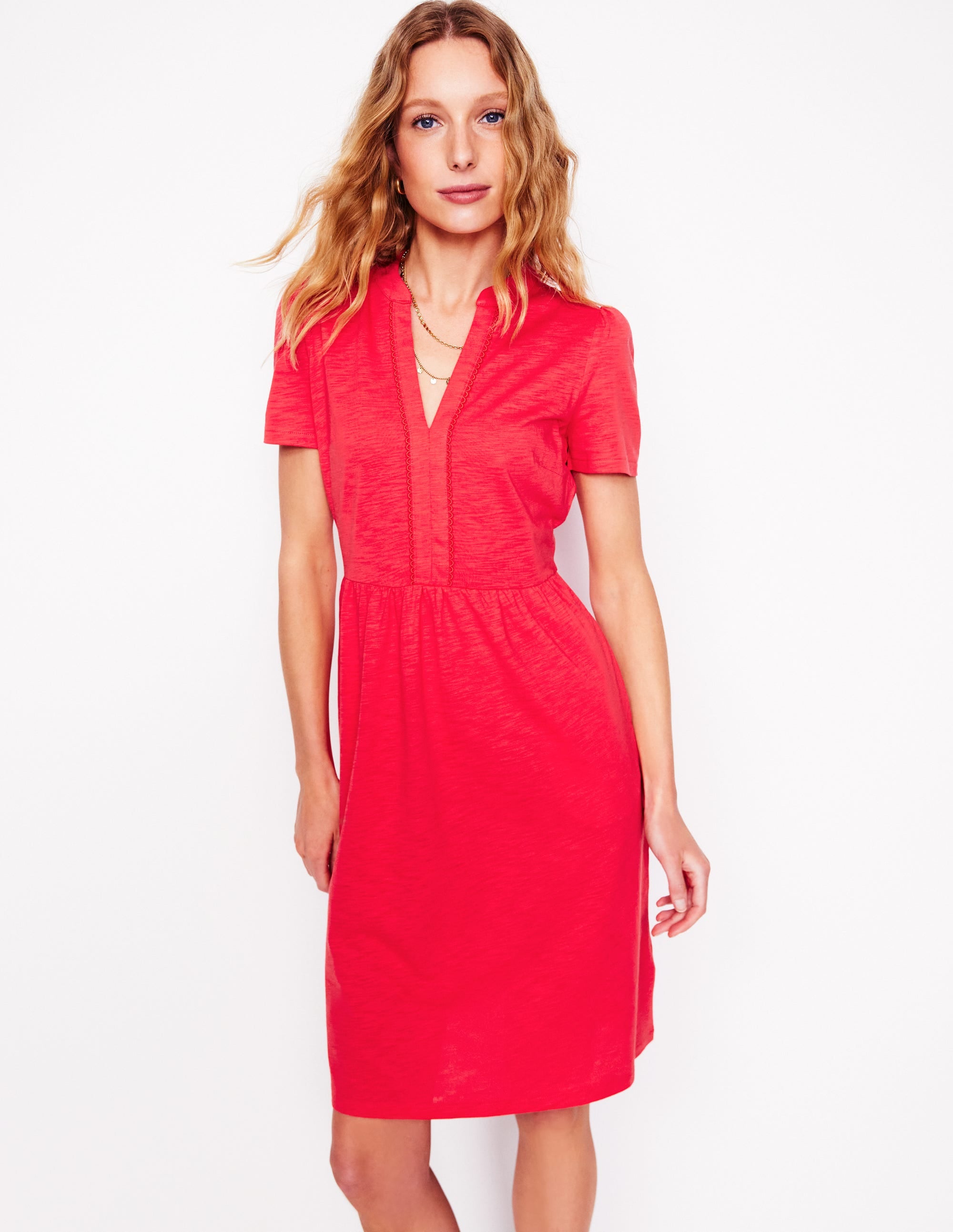 A woman wearing a bright red short jersey dress with short sleeves and a gathered waist, standing against a white background.