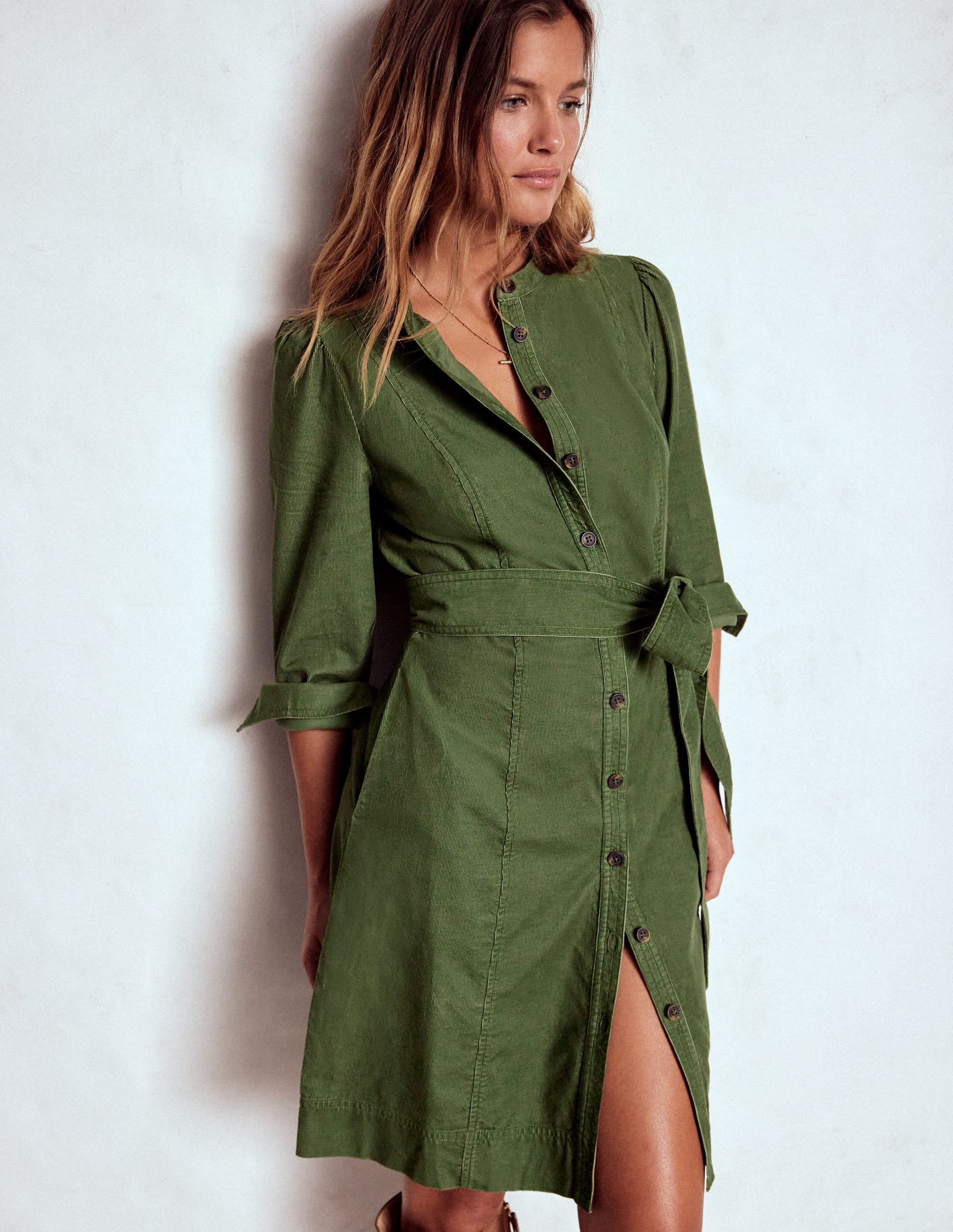 A woman wearing a green short dress with a fabric belt, blouson sleeves, and button-down front, standing against a plain background.