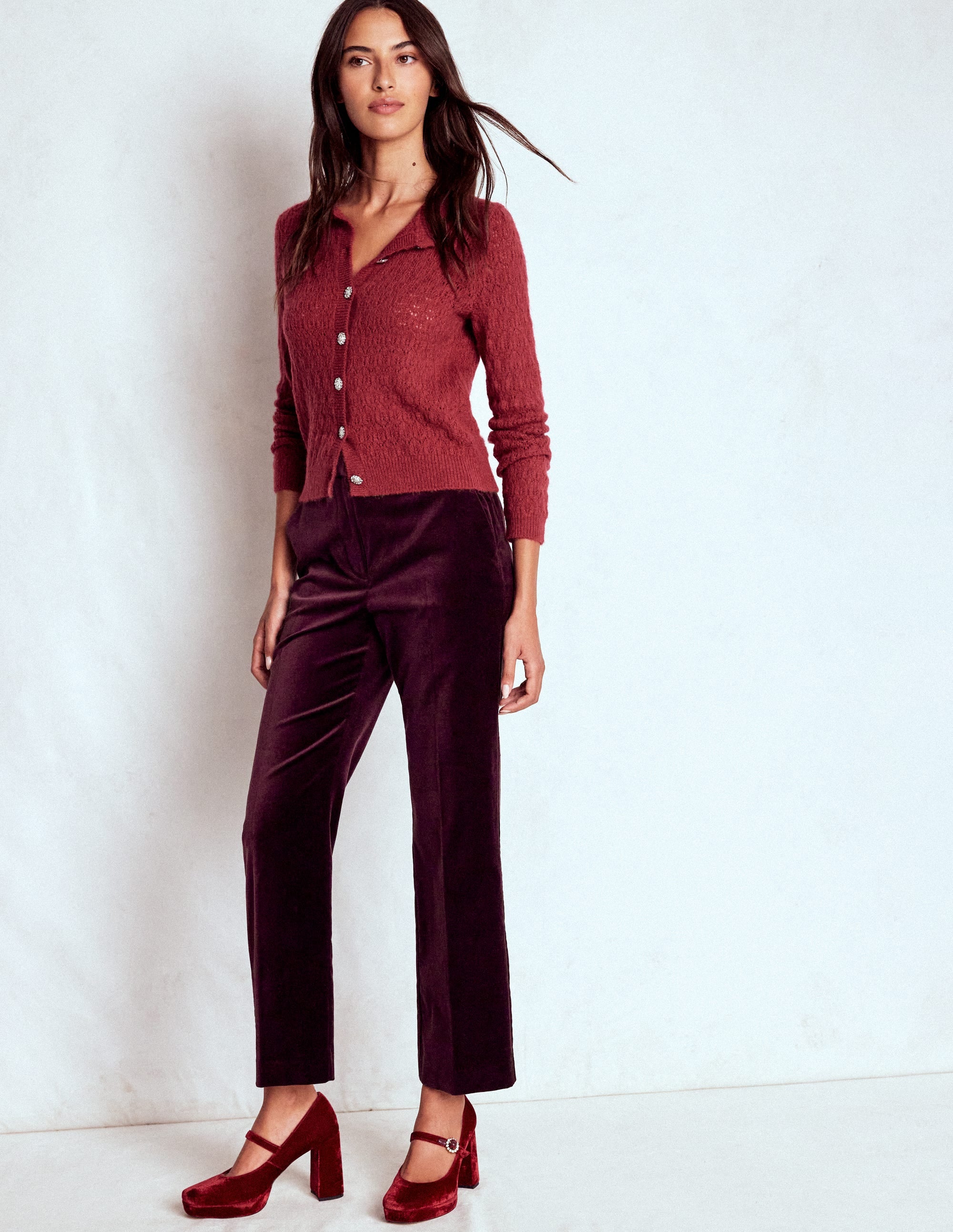 A woman wearing dark cherry velvet trousers with a tailored fit, natural waist, and functional pockets, paired with a red top and matching red heels.