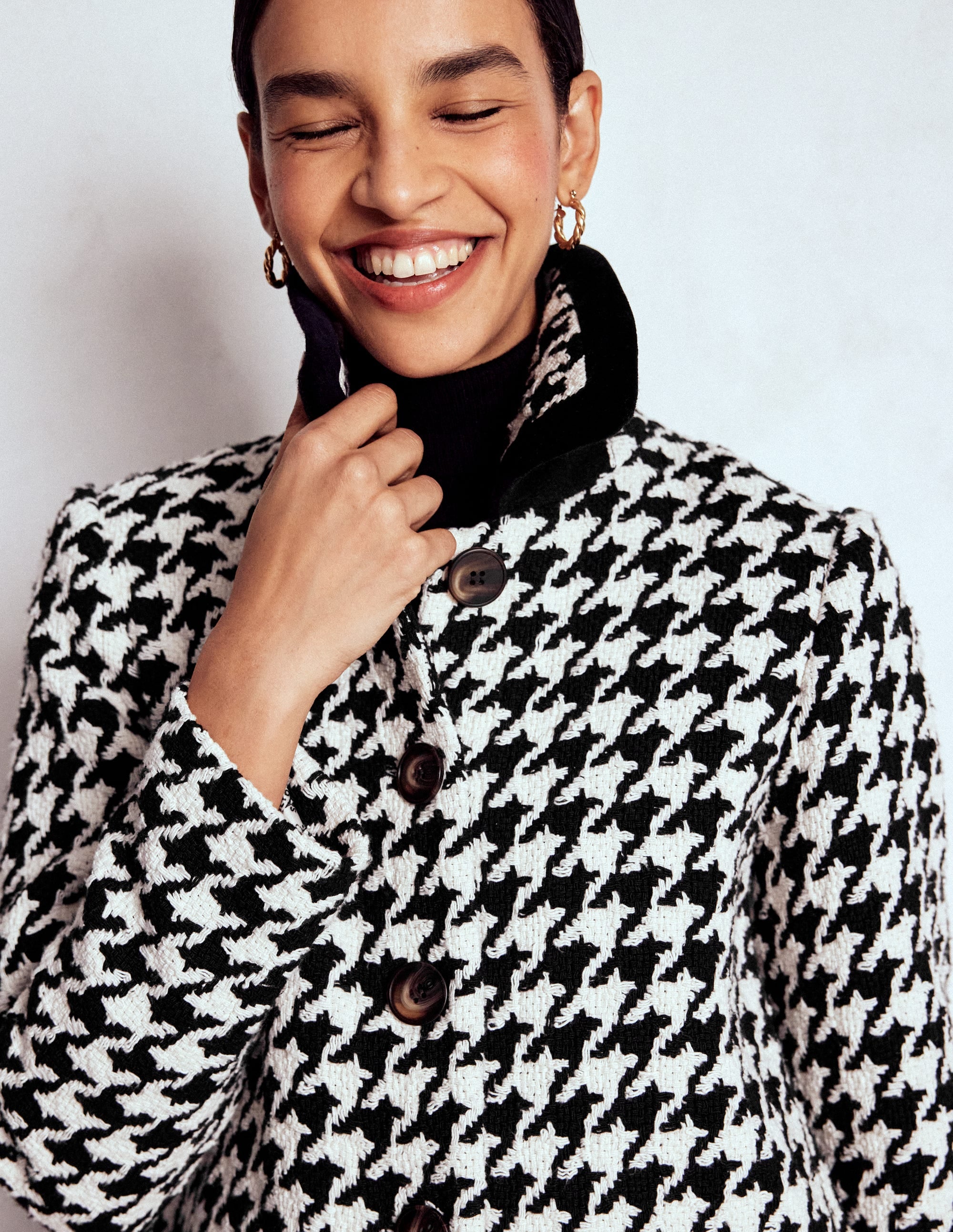 A woman wearing a knee-length black and white houndstooth swing coat with pleats at the back, button closure, and pockets, paired with gold hoop earrings.