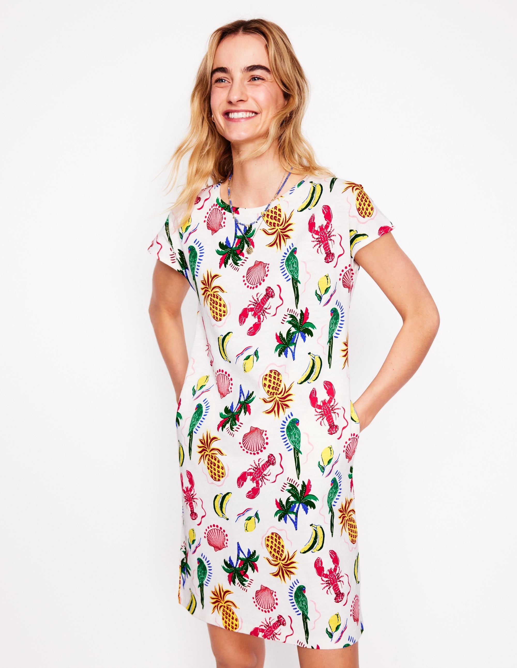 A woman wearing a knee-length white jersey T-shirt dress with a colorful tropical pattern including pineapples, lobsters, parrots, and palm trees.