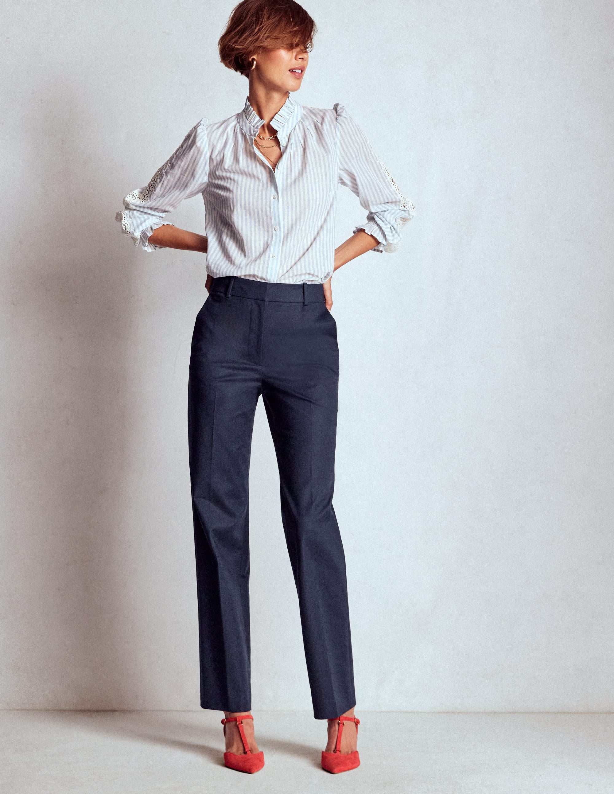 A woman wearing navy full-length trousers with a high waist, front crease, and a slightly tapered leg, paired with a striped blouse and red heels.