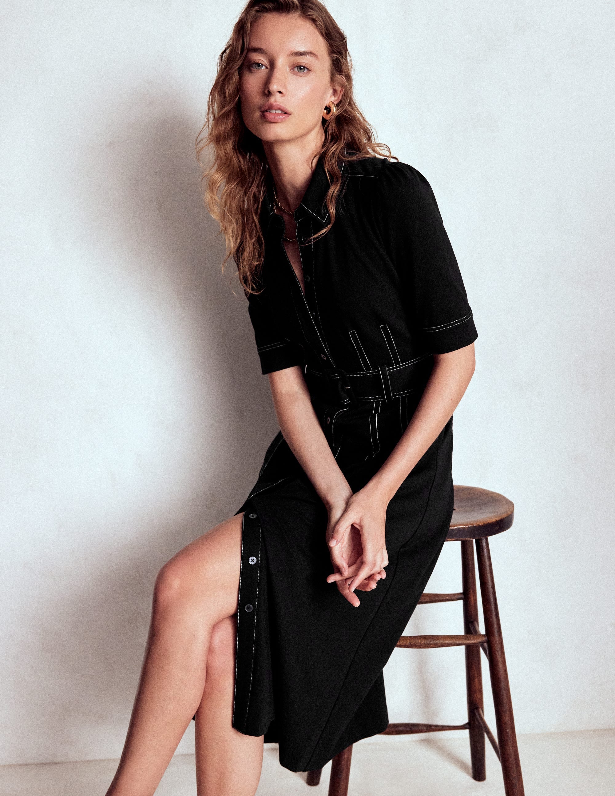 A woman wearing a black shirt dress with a collar, short sleeves, a wide belt, and pockets, sitting on a wooden stool.