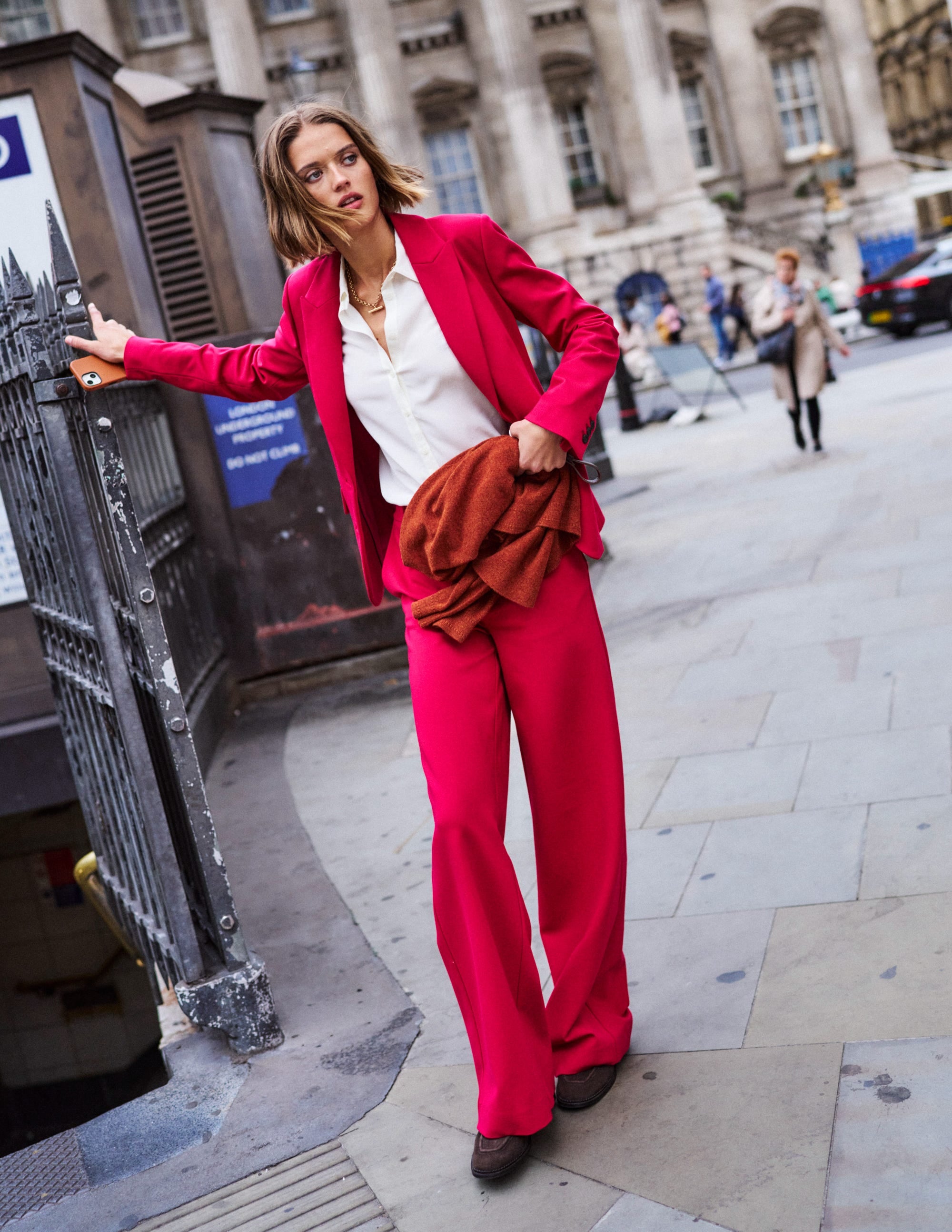 A woman wearing bright red wide-leg trousers, a white shirt, and a red blazer, standing on a city street with historic buildings in the background.