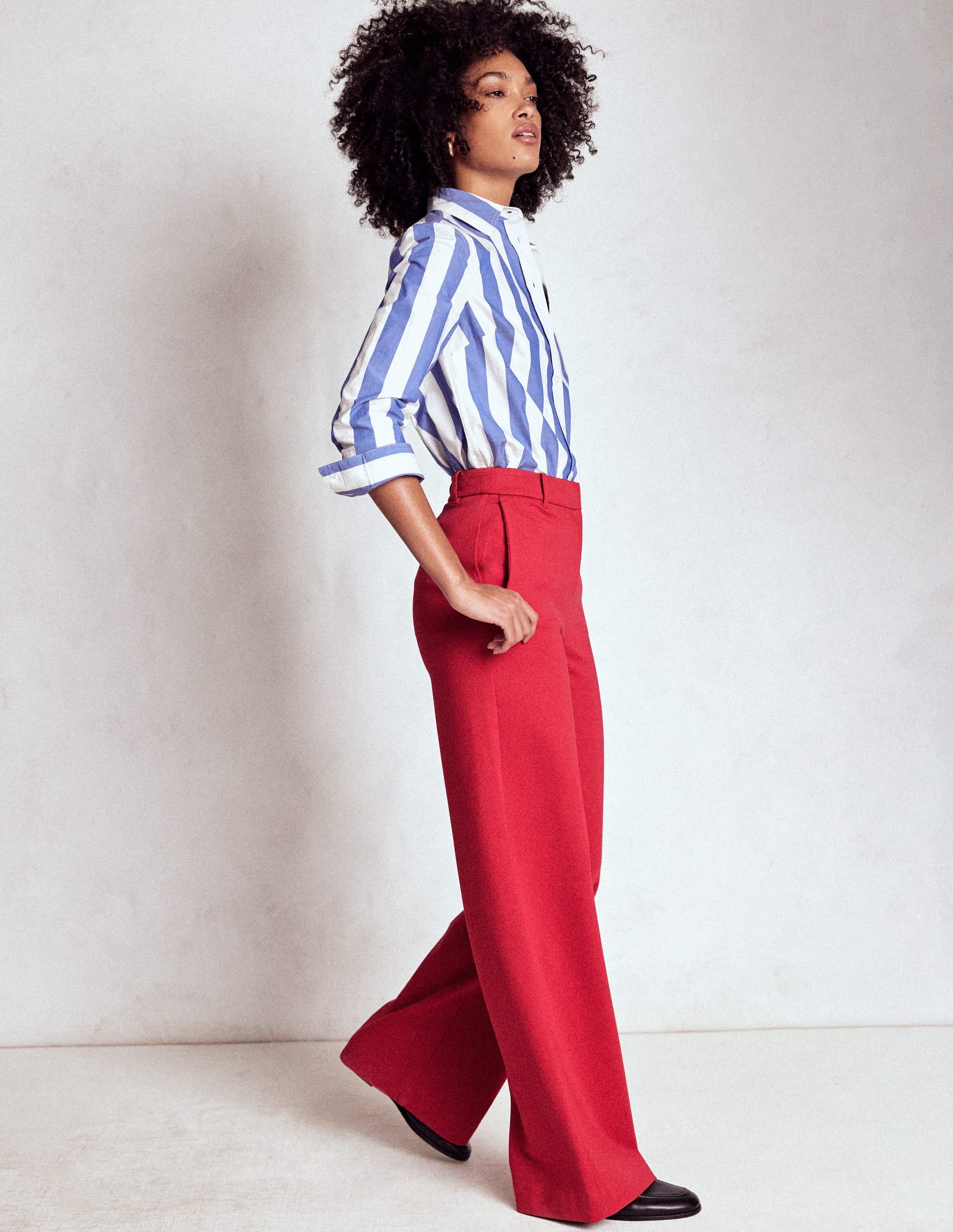 A woman wearing high-waisted, Carmine Red trousers with a subtly flared cut, functional pockets, and a concealed hook and bar fastening, paired with a blue and white striped shirt.