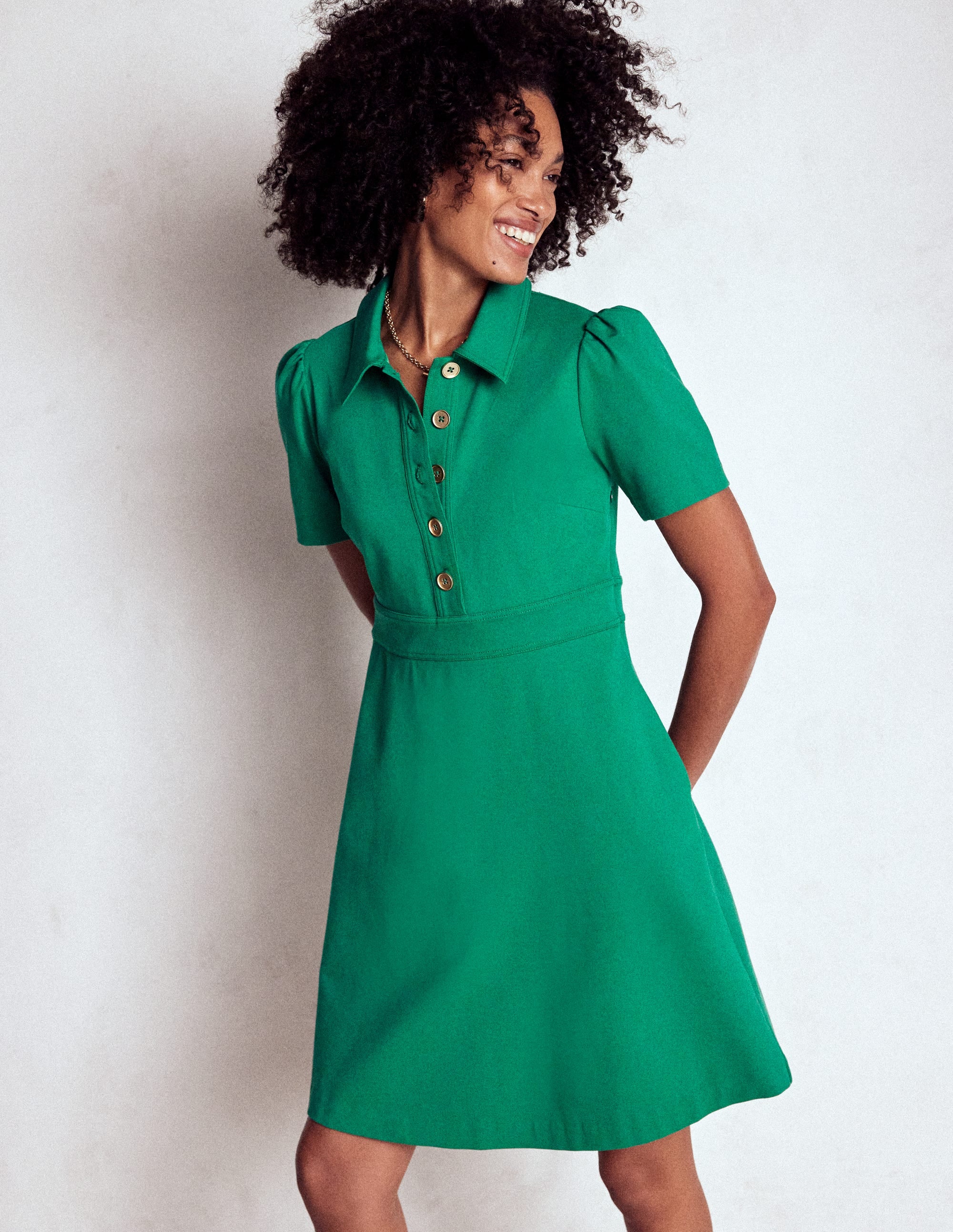 A woman wearing a vibrant green short dress with short puffed sleeves, a button-up front with gold buttons, and side seam pockets, standing against a plain background.