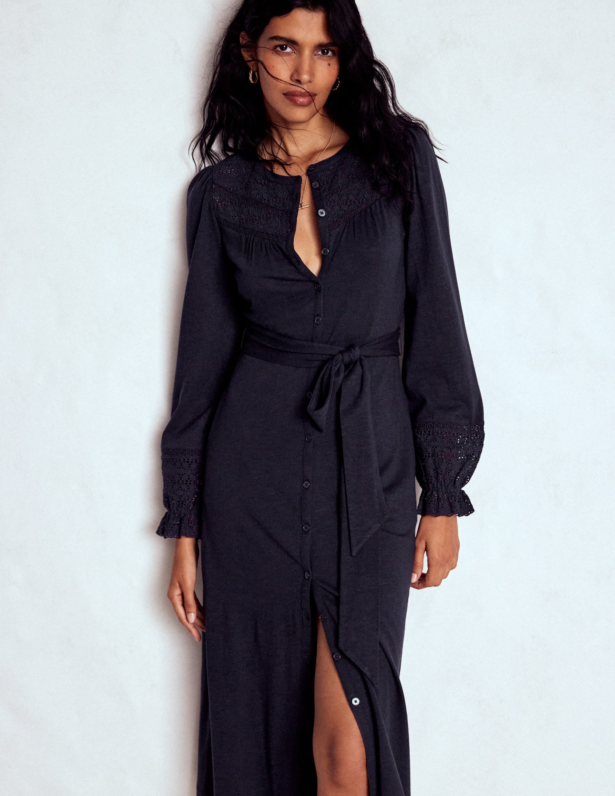 A woman wearing a navy dress with broderie detailing, long sleeves with ruffled cuffs, a button-down front, and a waist tie.