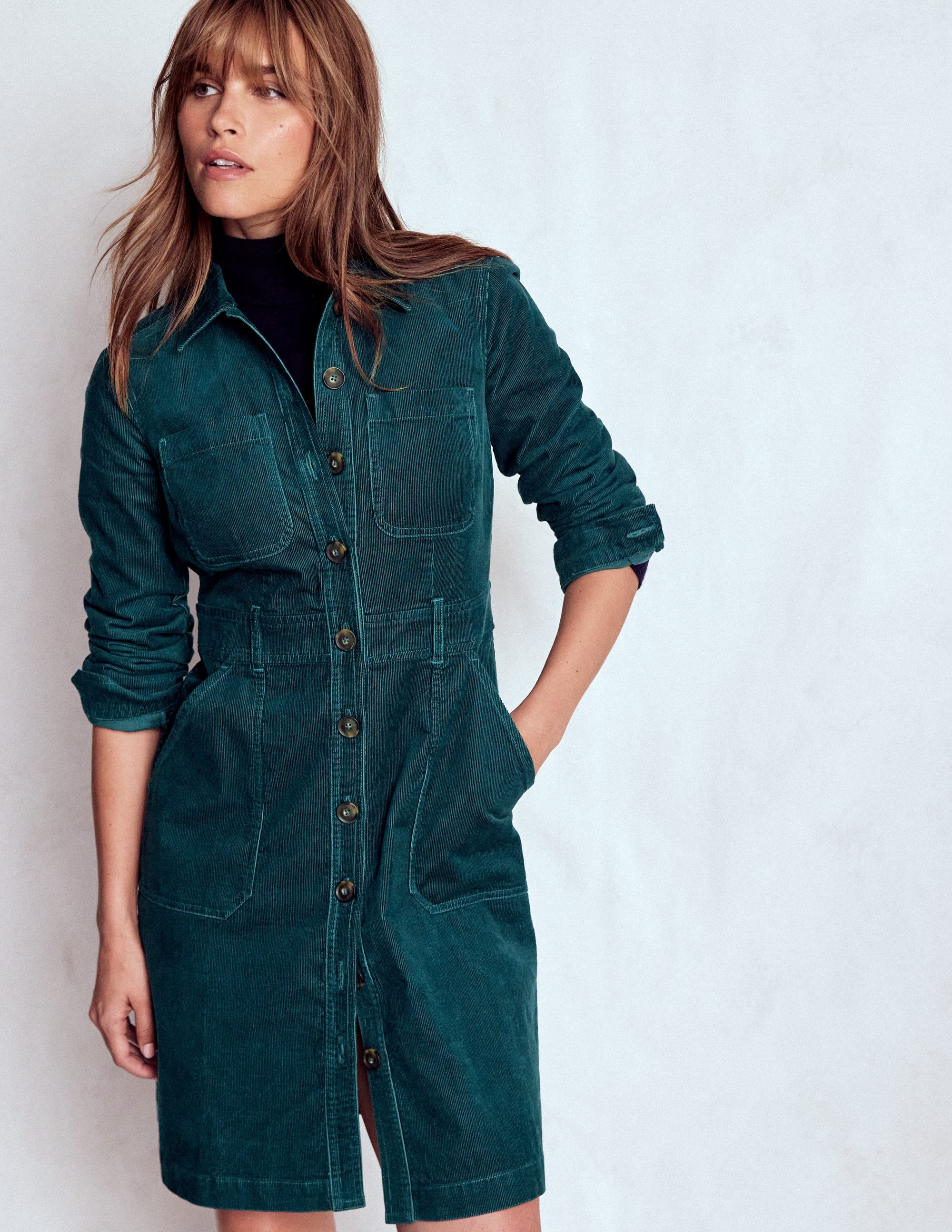 A woman wearing a midnight teal fitted short dress made of cotton cord fabric with utility details, including a button-down front and chest pockets.