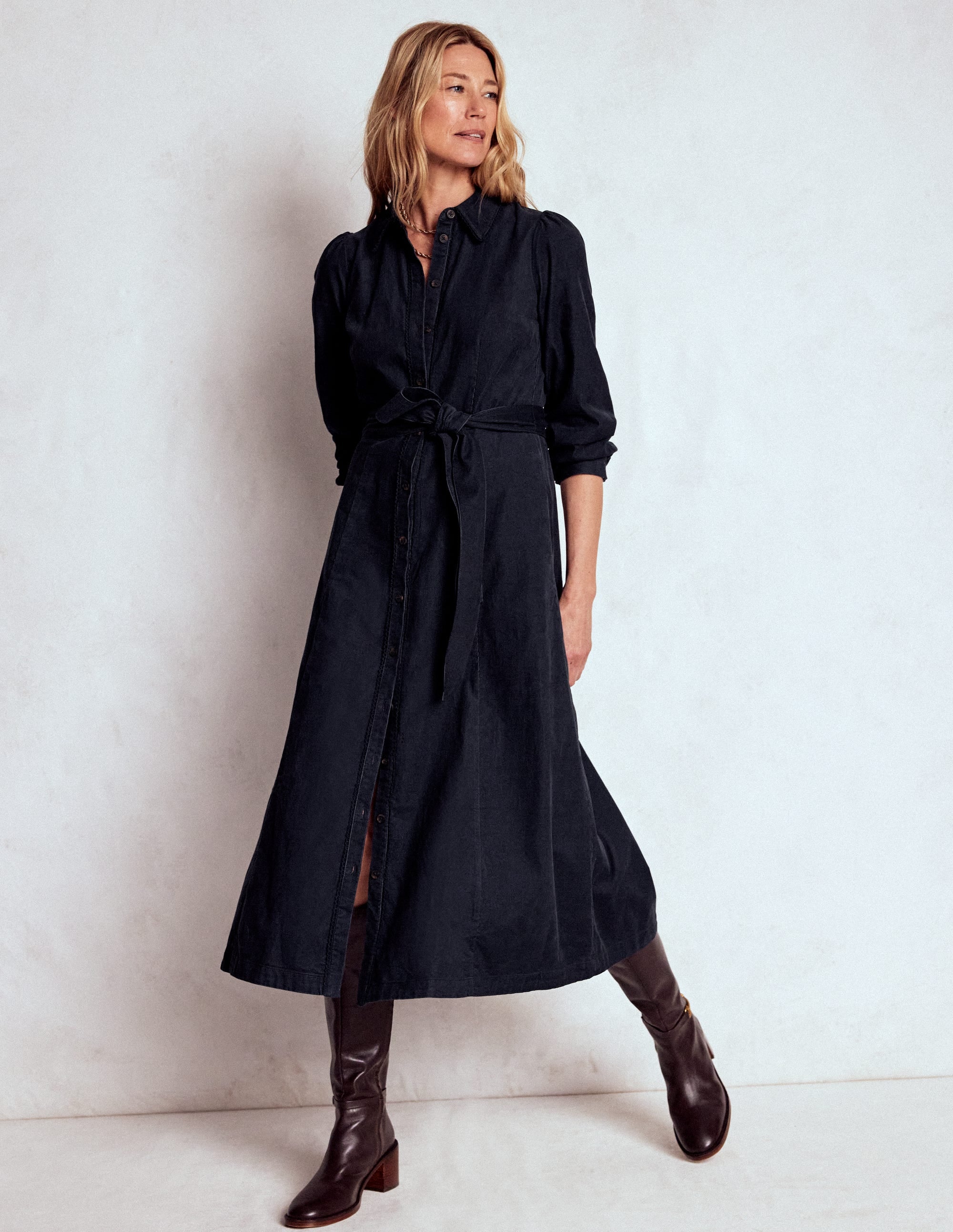 A woman wearing a midnight blue midi dress with statement sleeves, a side belt, and pockets, paired with brown knee-high boots.