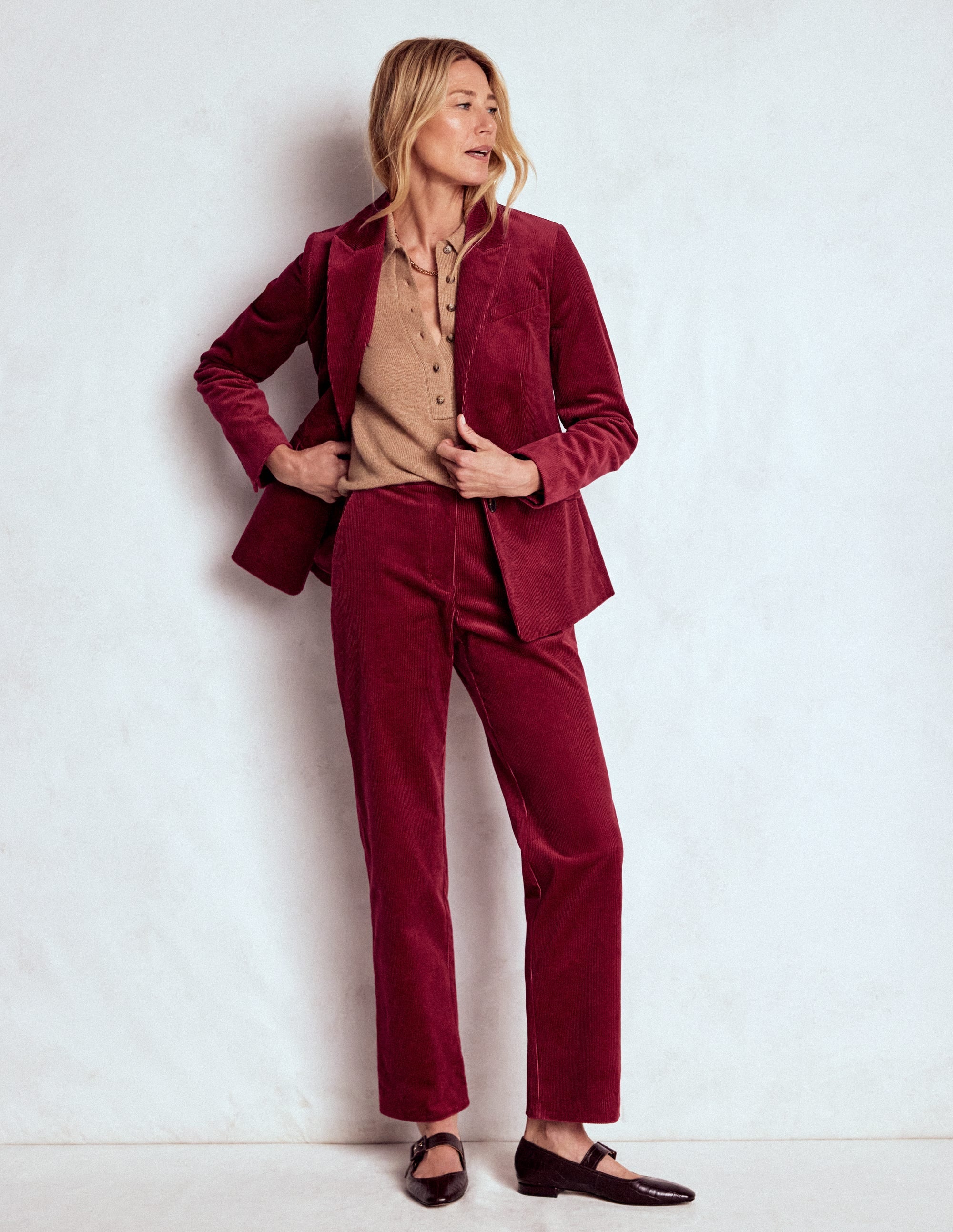 A woman wearing burgundy red cord trousers, a matching blazer, and a beige top, standing against a plain background.