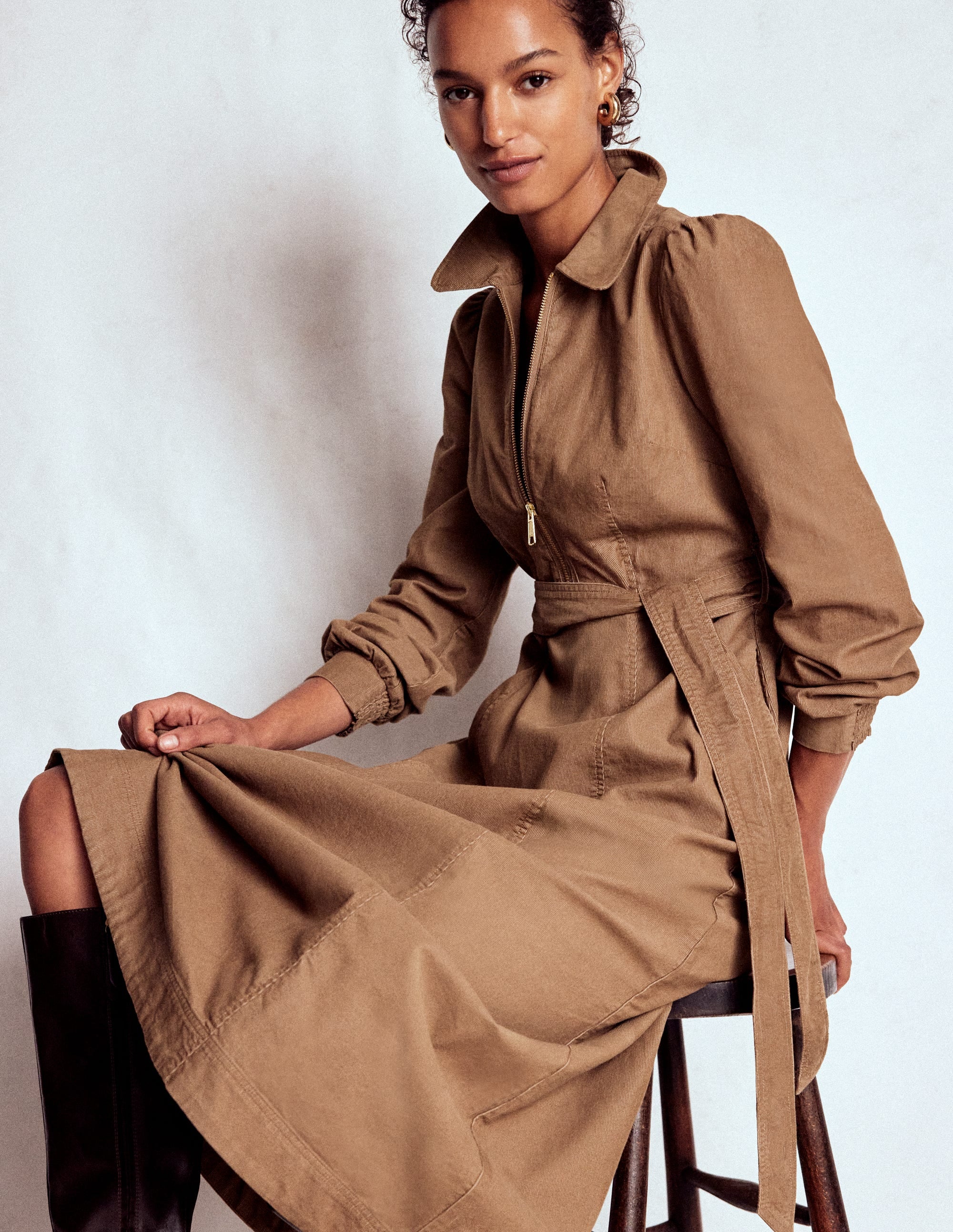 A woman wearing a beige midi dress with a zip-up top, fabric belt, and side seam pockets, sitting on a stool.
