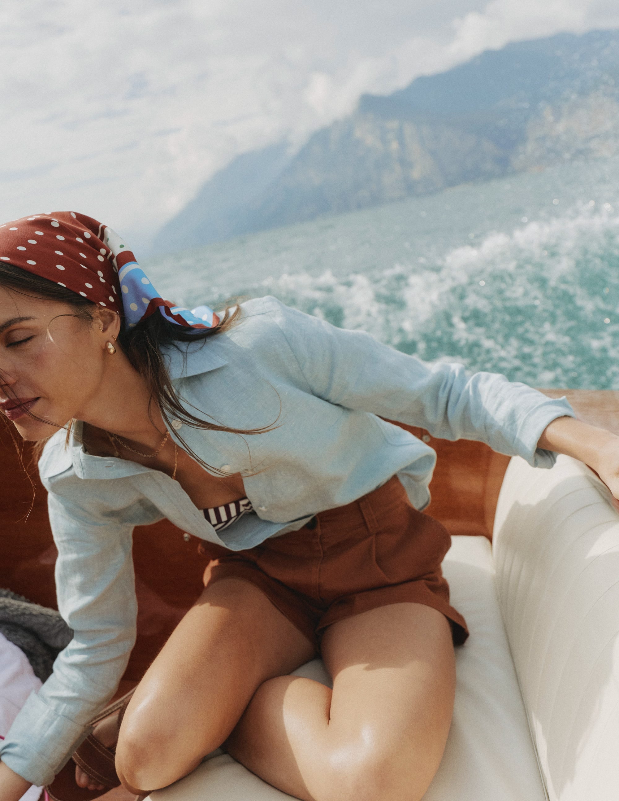 A woman wearing a light blue linen shirt with a collar and buttons, sitting on a boat with water and mountains in the background, wearing a red polka dot headscarf.