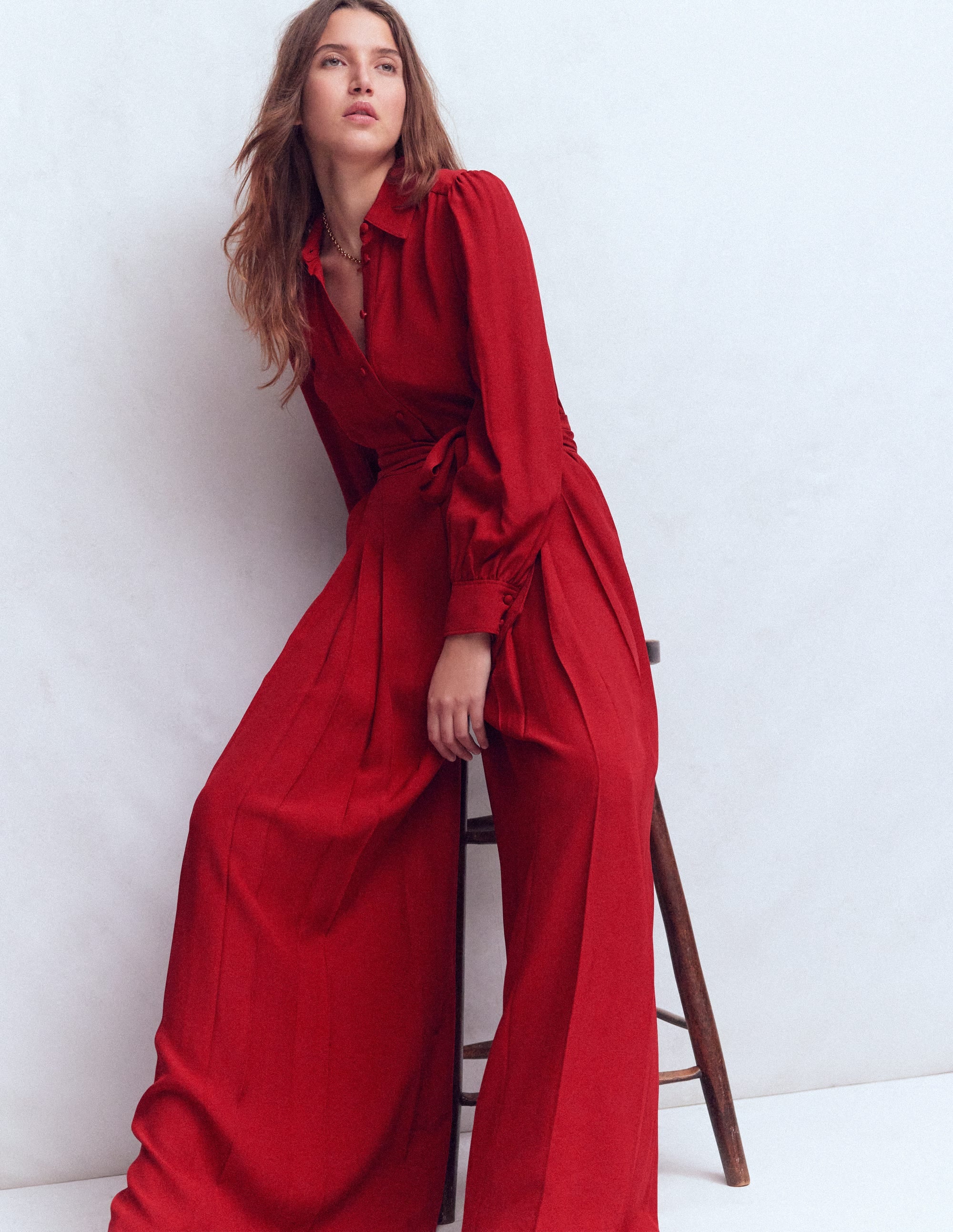 A woman wearing a crimson red button-up palazzo jumpsuit with wide legs, soft volume sleeves, and a self-fabric belt, standing against a plain background.