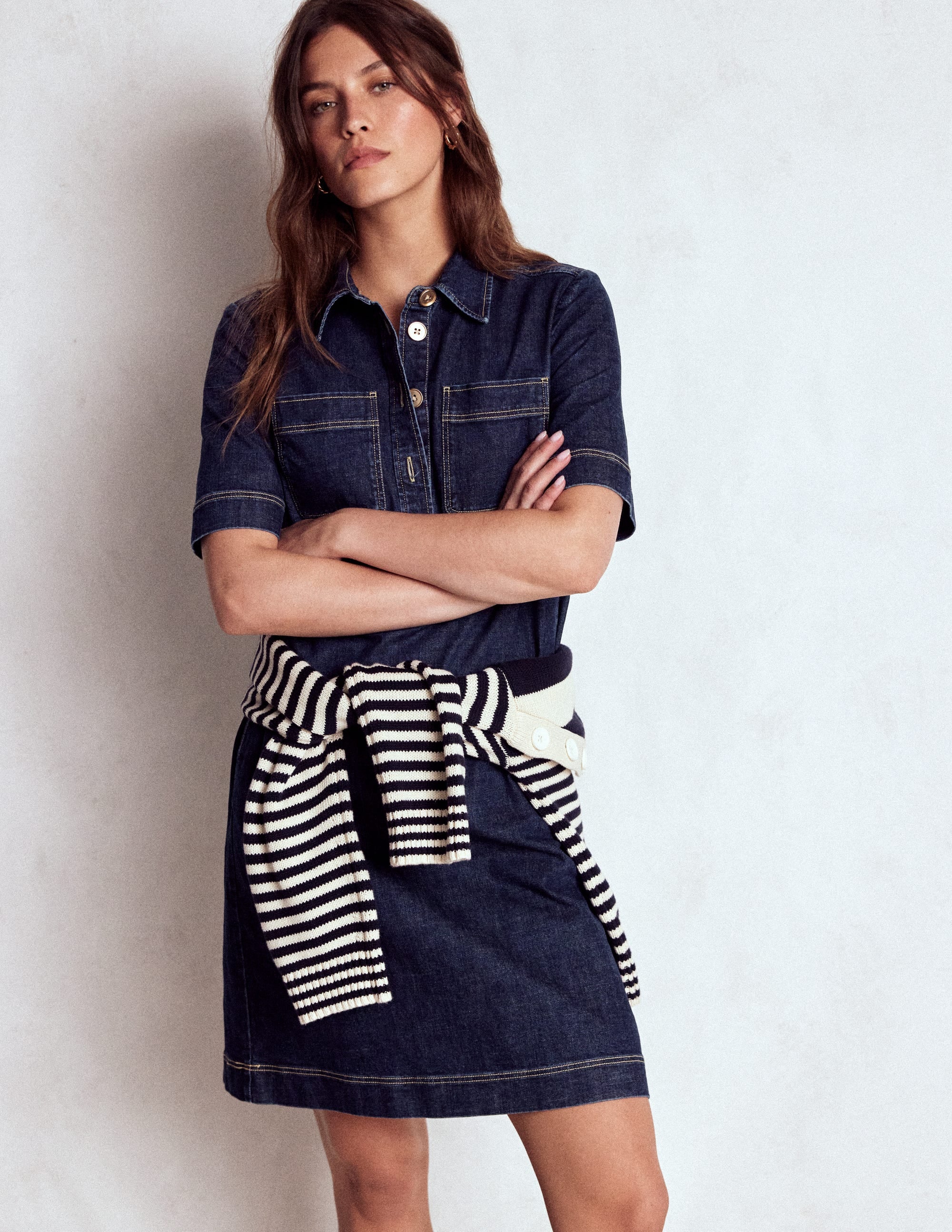 A woman wearing a dark indigo denim shift dress with short sleeves, a collar, and side pockets, standing against a plain background.