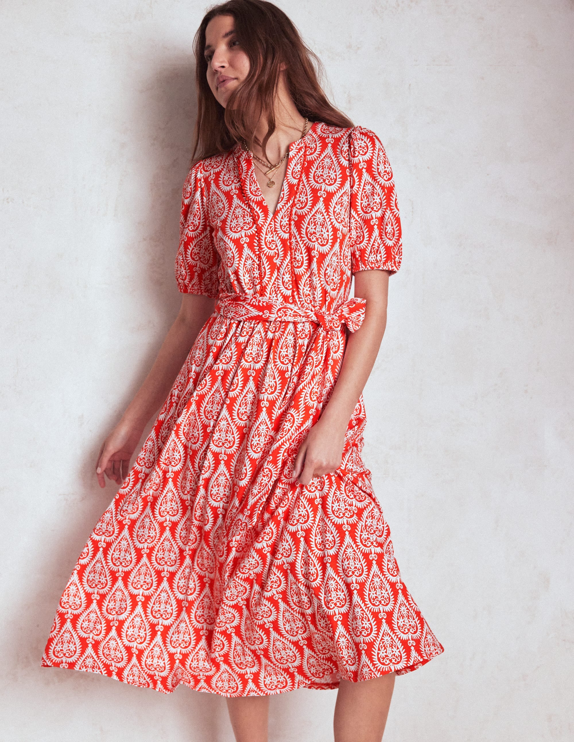 A woman wearing a red midi dress with white ornate geometric patterns, puff sleeves, scallop trim, and a self-fabric belt, standing against a plain background.