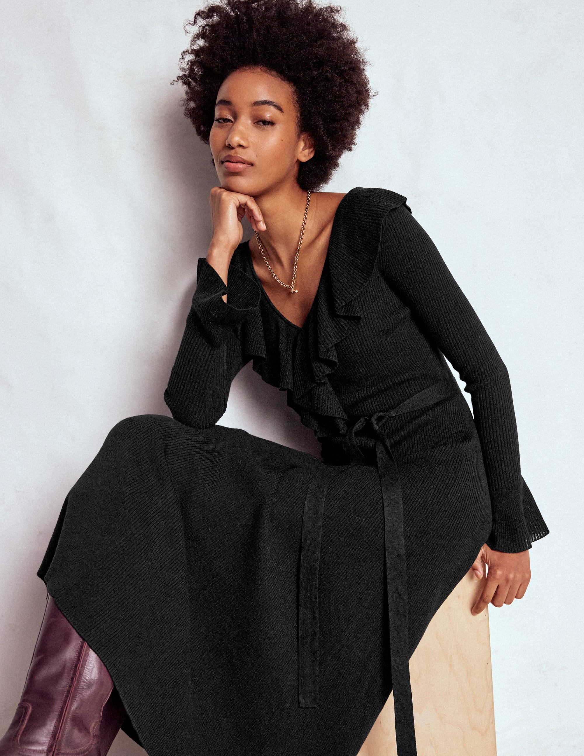 A woman wearing a black knitted maxi dress with ruffled and frilled sleeves, sitting against a plain background.